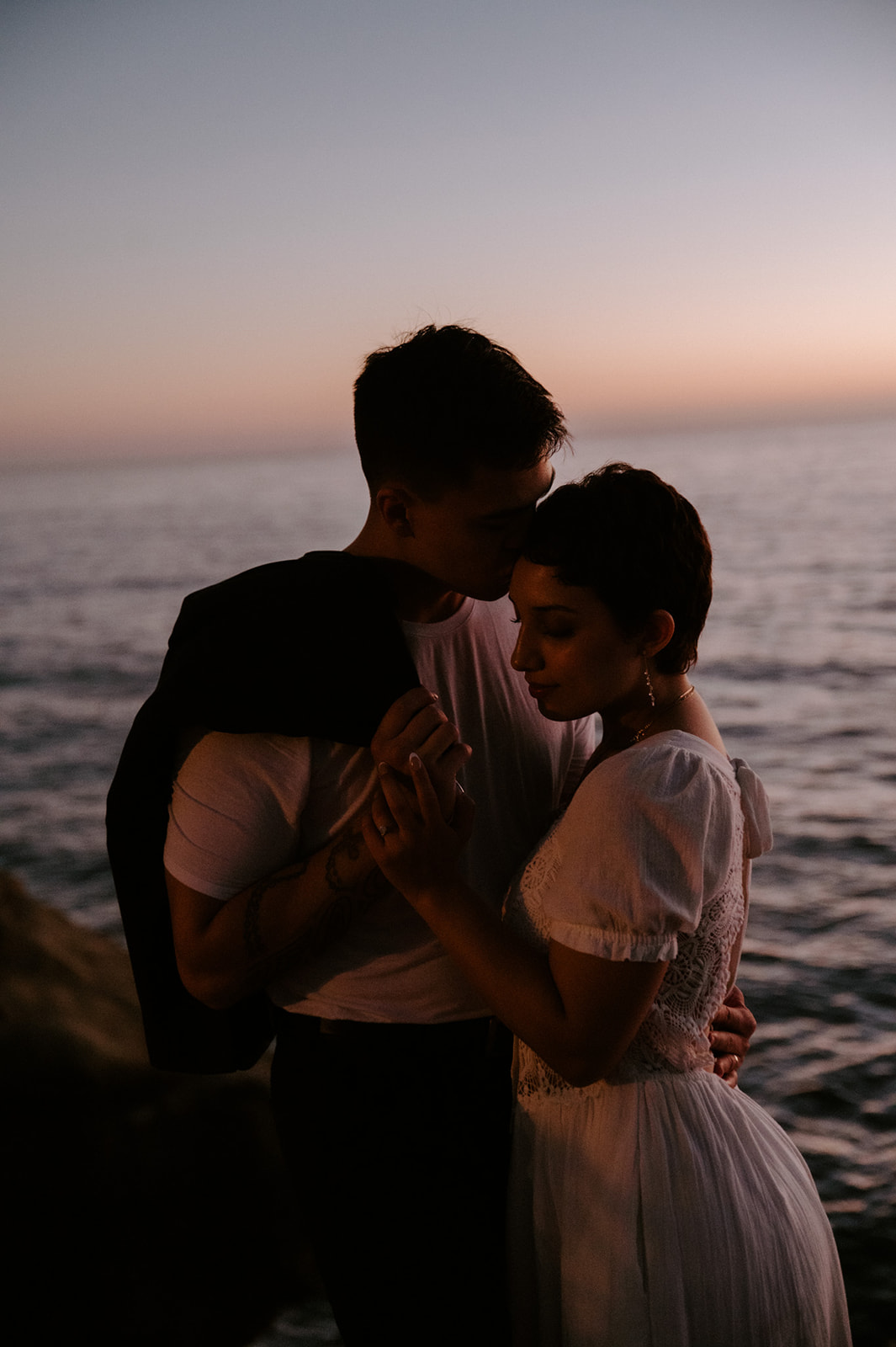 Groom kissing bride’s forehead as they hold each other close by the ocean at sunset, a calm and intimate elopement checklist moment.
