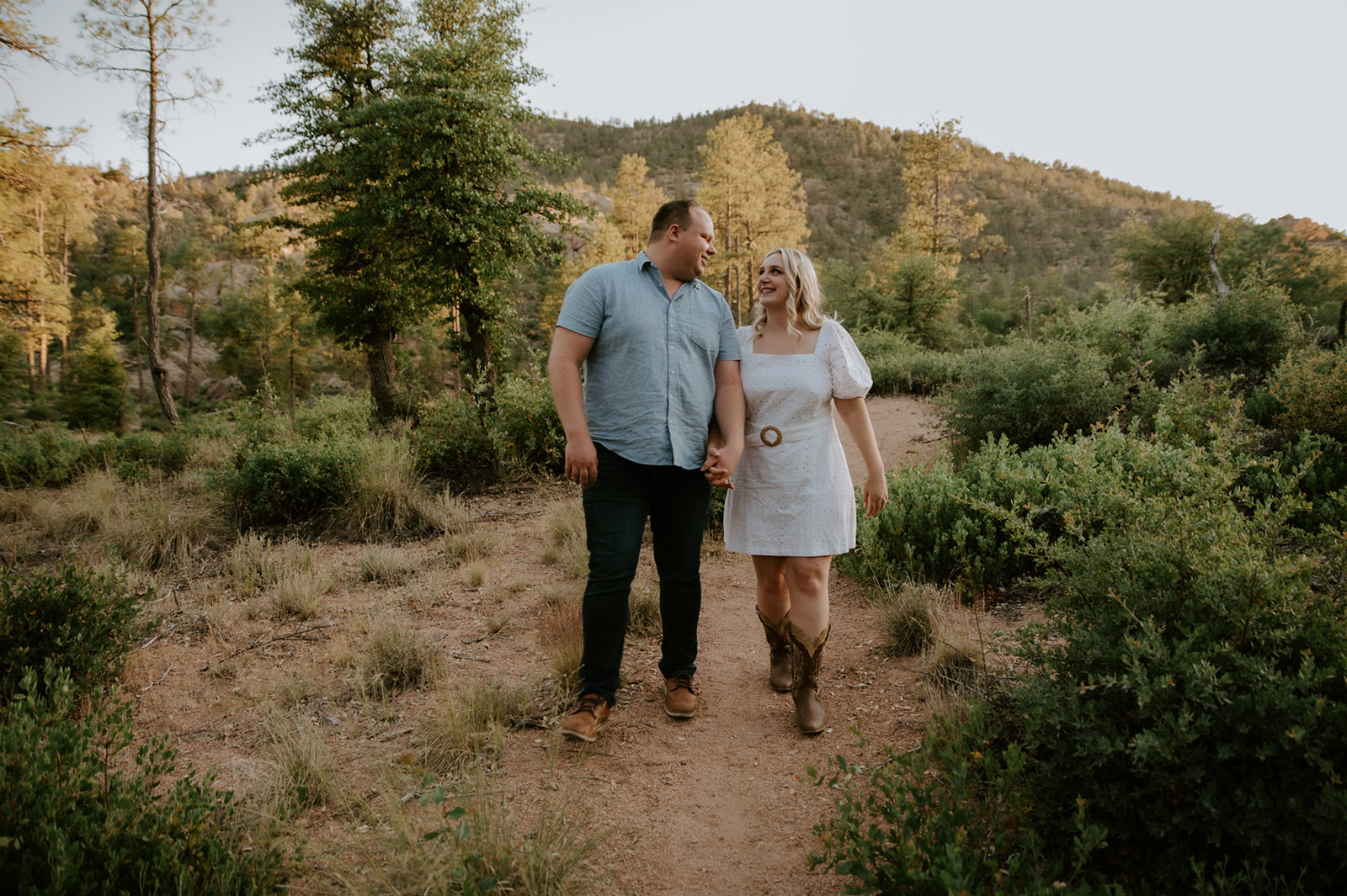 Couple walking hand in hand along a trail surrounded by greenery and mountain views
