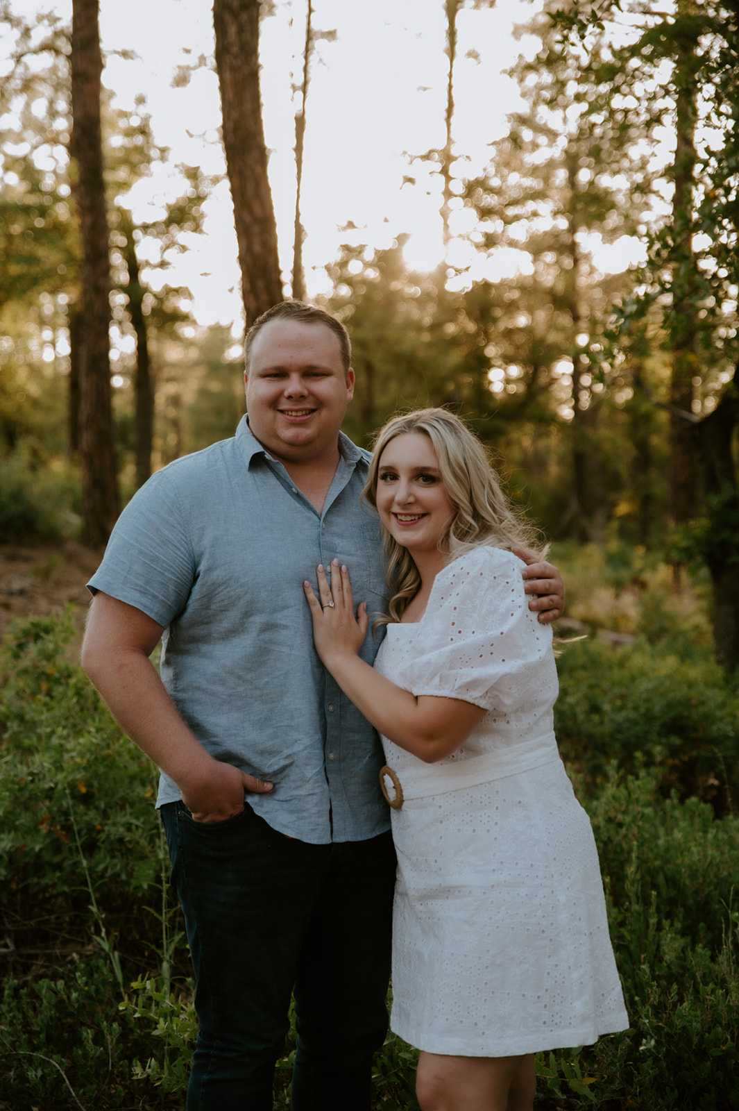 Couple smiling at the camera while standing together in a sunlit pine forest in Flagstaff
