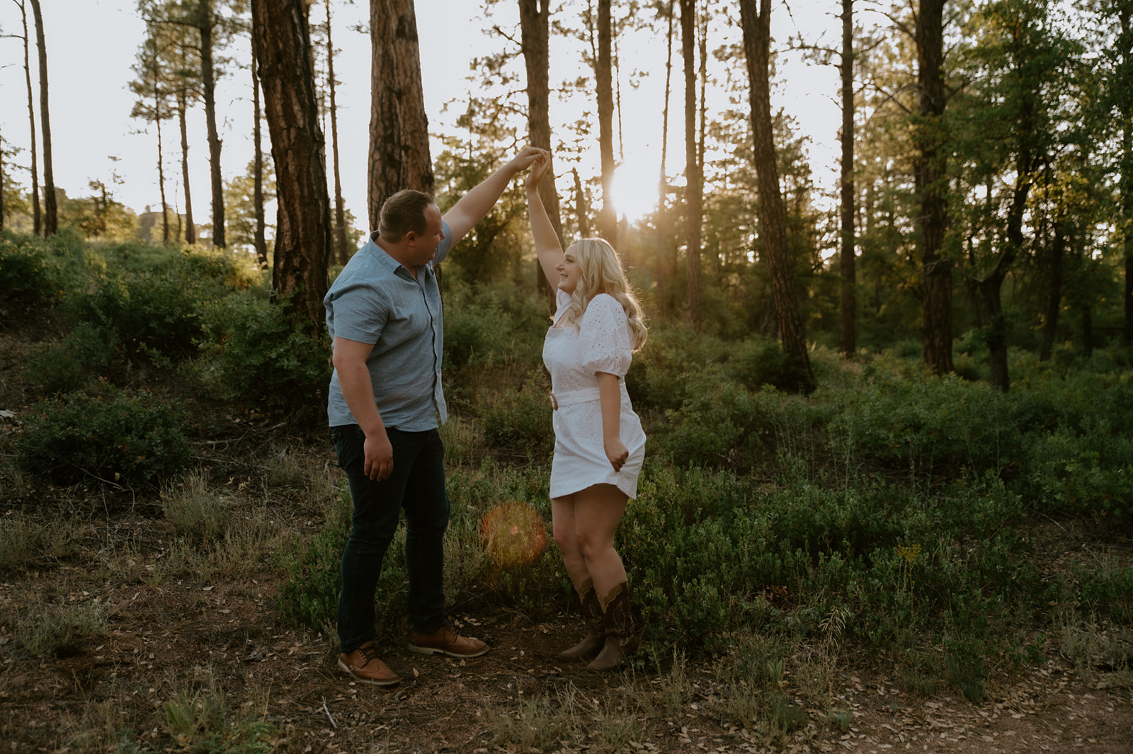 Couple twirling together and laughing in a sunlit forest clearing during golden hour
