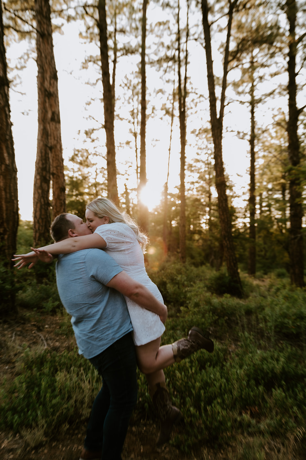Groom lifting bride into the air for a playful kiss in a sunlit pine forest during golden hour
