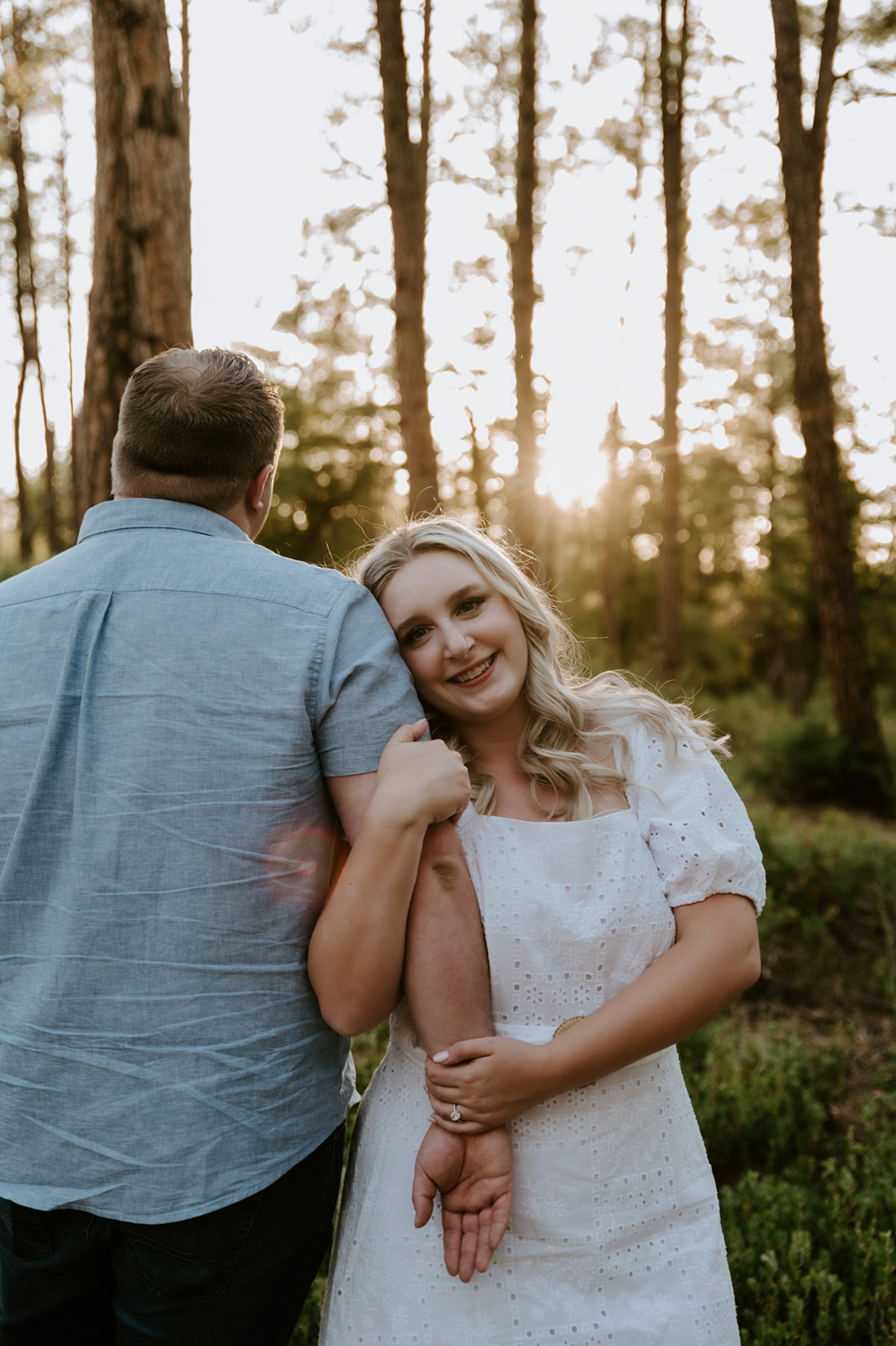 Bride leaning on her partner’s shoulder and smiling in a dreamy forest setting at sunset
