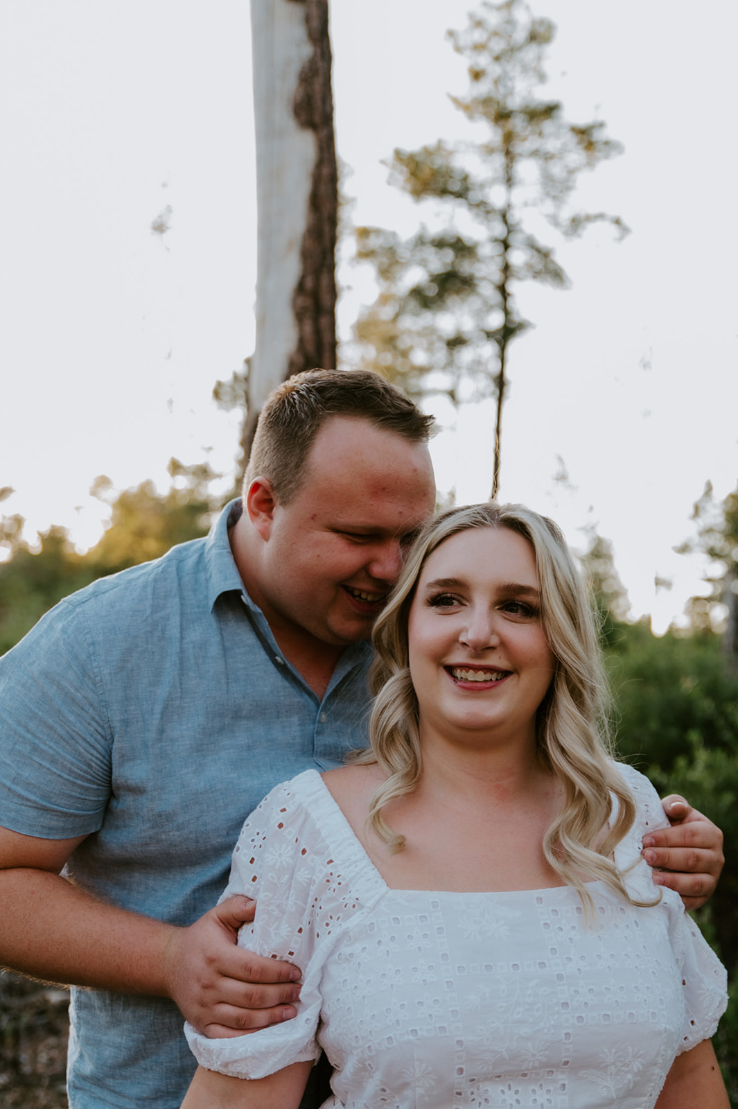 Groom hugging bride from behind as she smiles in a soft, glowing forest scene
