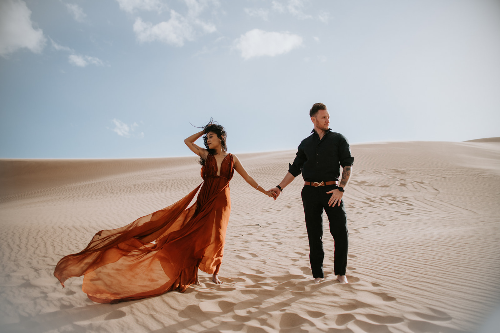 Couple holding hands and walking across wide open dunes, flowing dress adding movement to the landscape
