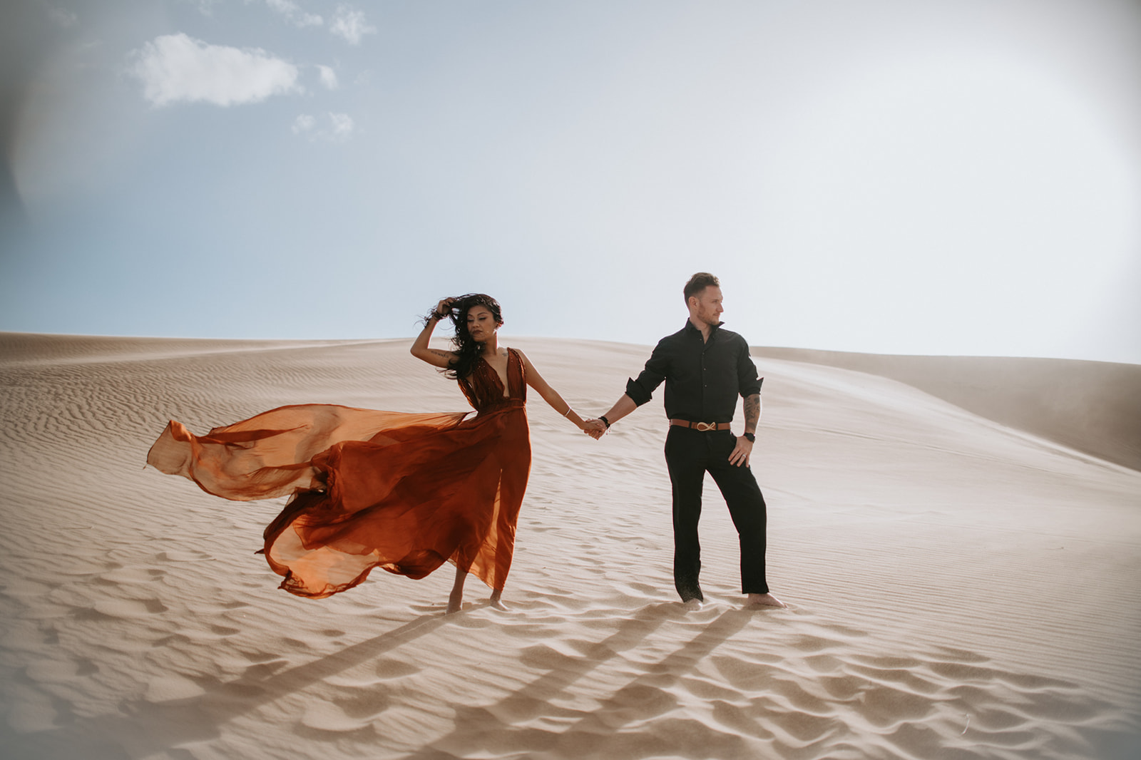 Couple walking hand in hand across wind-swept sand dunes, red dress flowing in the breeze during an engagement session
