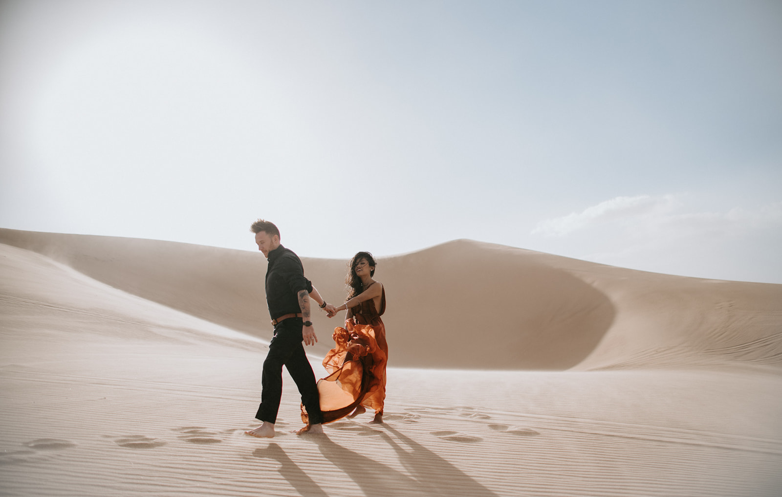 Couple walking hand in hand across open dunes, soft light and long shadows creating depth
