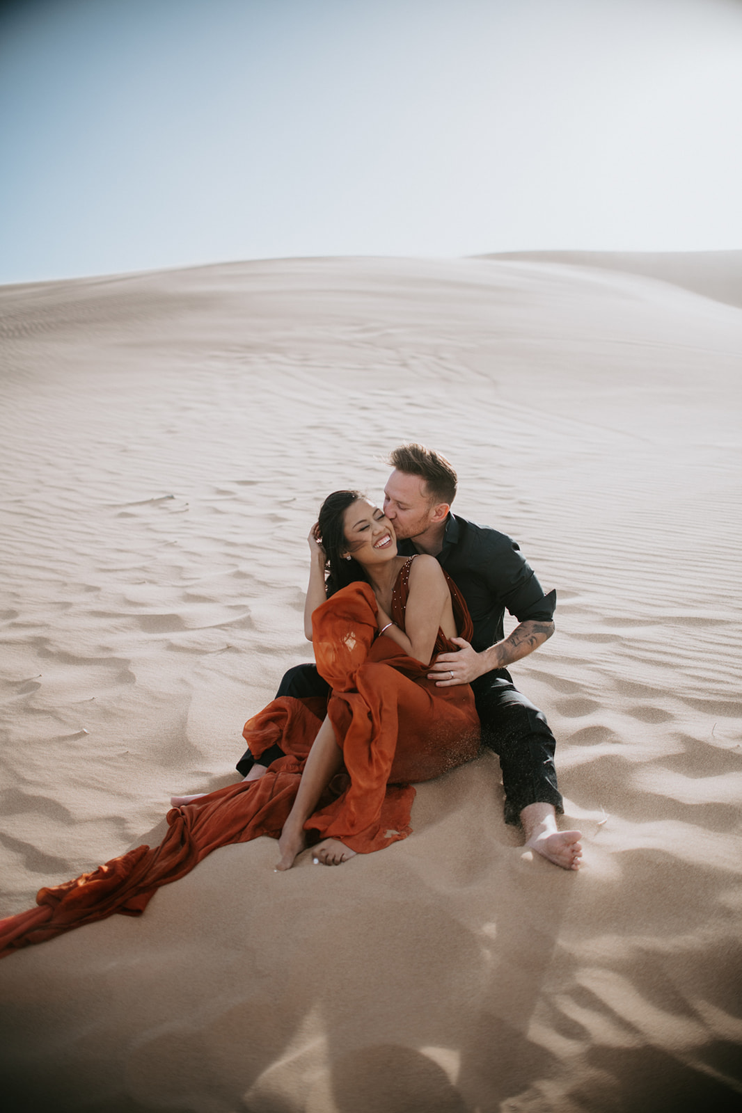 Couple sitting close together on sunlit dunes, warm tones and soft shadows creating a relaxed desert scene
