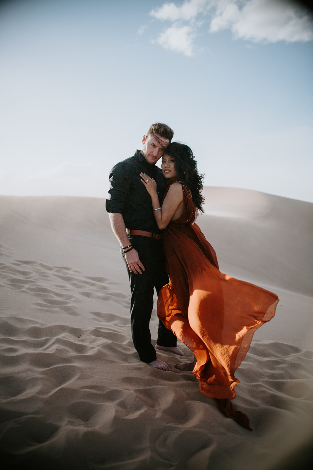 Couple standing together on a dune, wind moving through her dress during a sand dunes photoshoot
