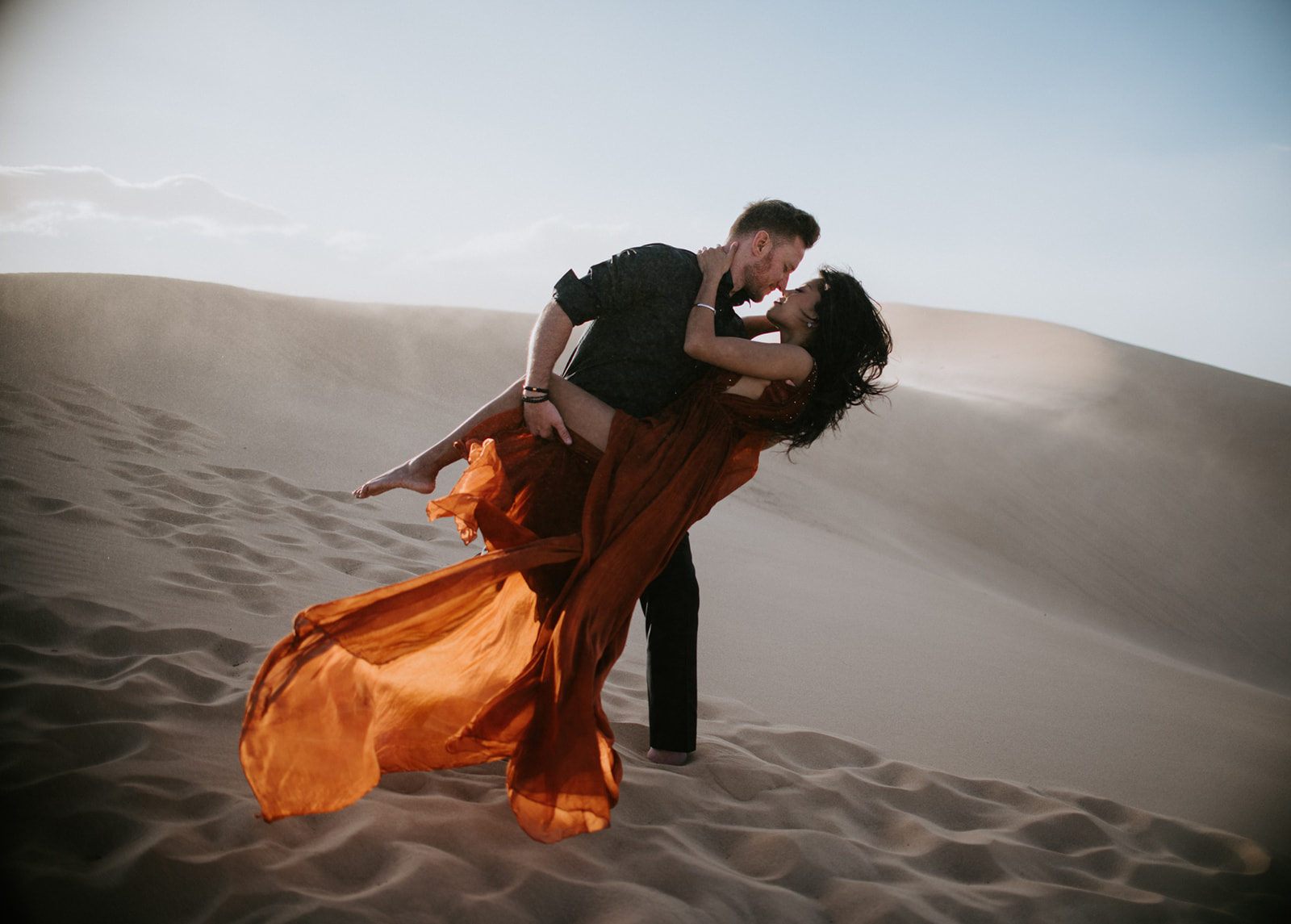 Couple mid-spin on a dune, red dress billowing in the wind during a playful sand dunes photoshoot
