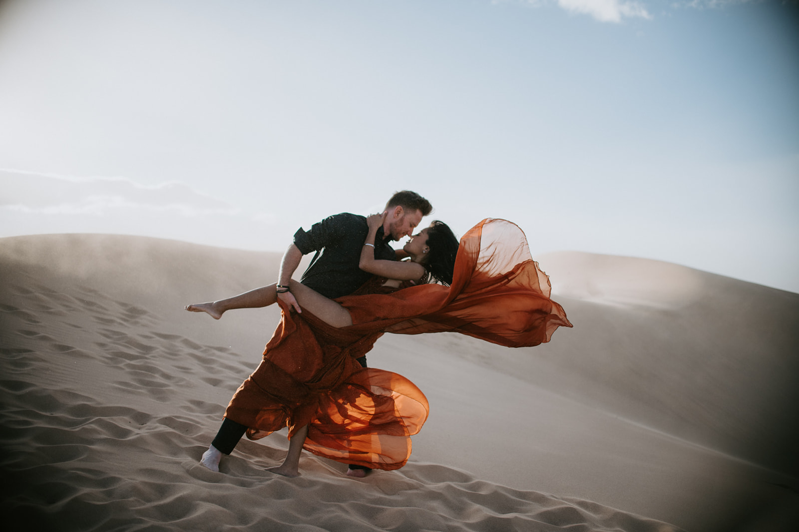 Couple lifting and spinning in the sand, dress moving dramatically in the wind during a sand dunes photoshoot
