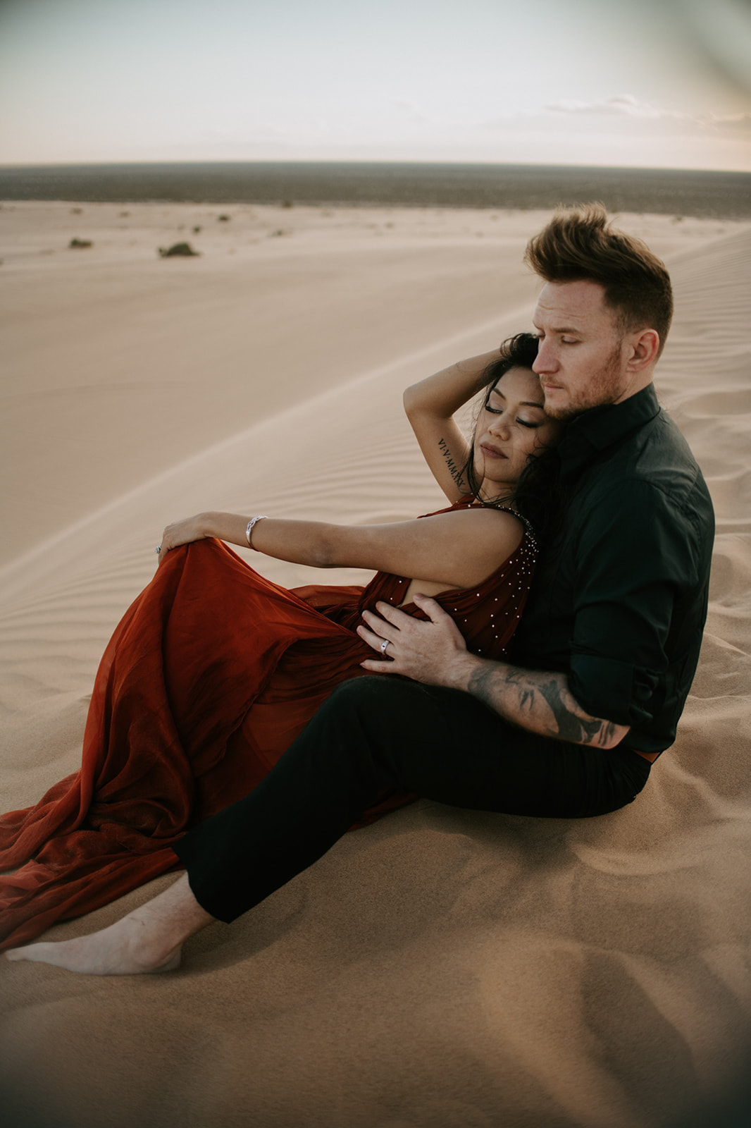Couple embracing closely on the dunes, wind catching her dress during a soft, intimate sand dunes photoshoot
