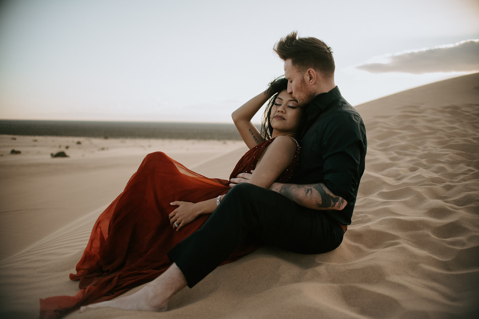 Couple sitting together on soft desert dunes, wrapped in a flowing red dress during a sand dunes photoshoot at sunset
