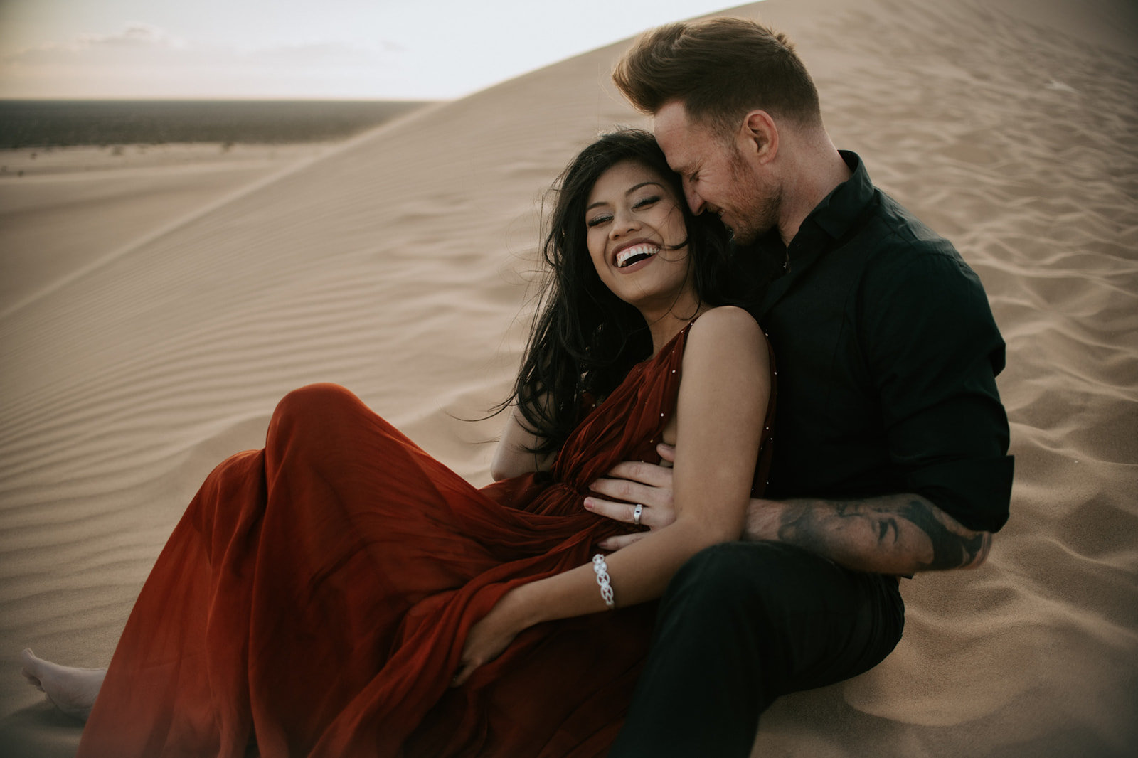 Couple sitting together on textured sand dunes, laughing and relaxed during an intimate sand dunes photoshoot