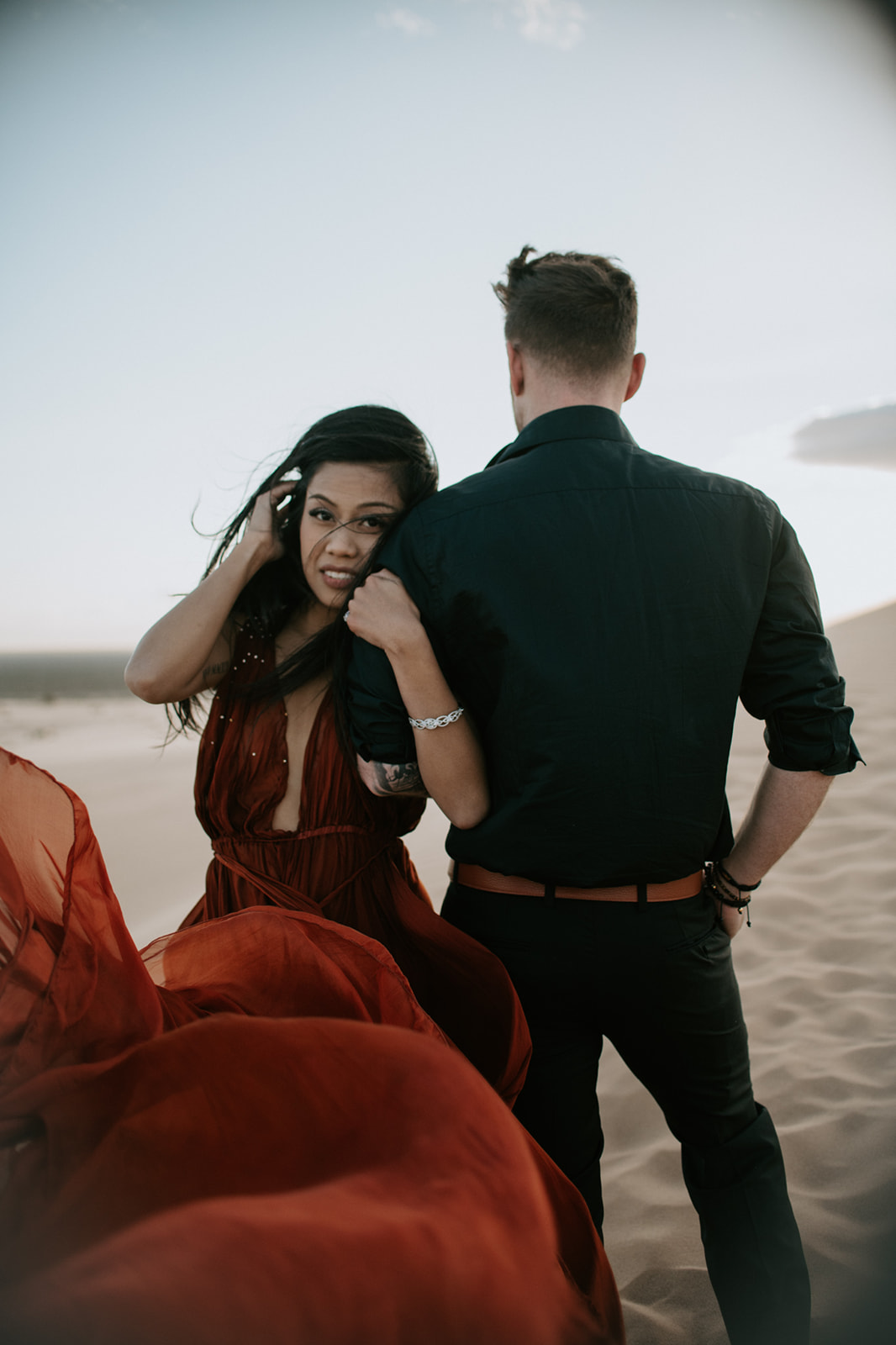 Couple walking along a dune ridge, wind catching the red dress and adding movement to the desert scene

