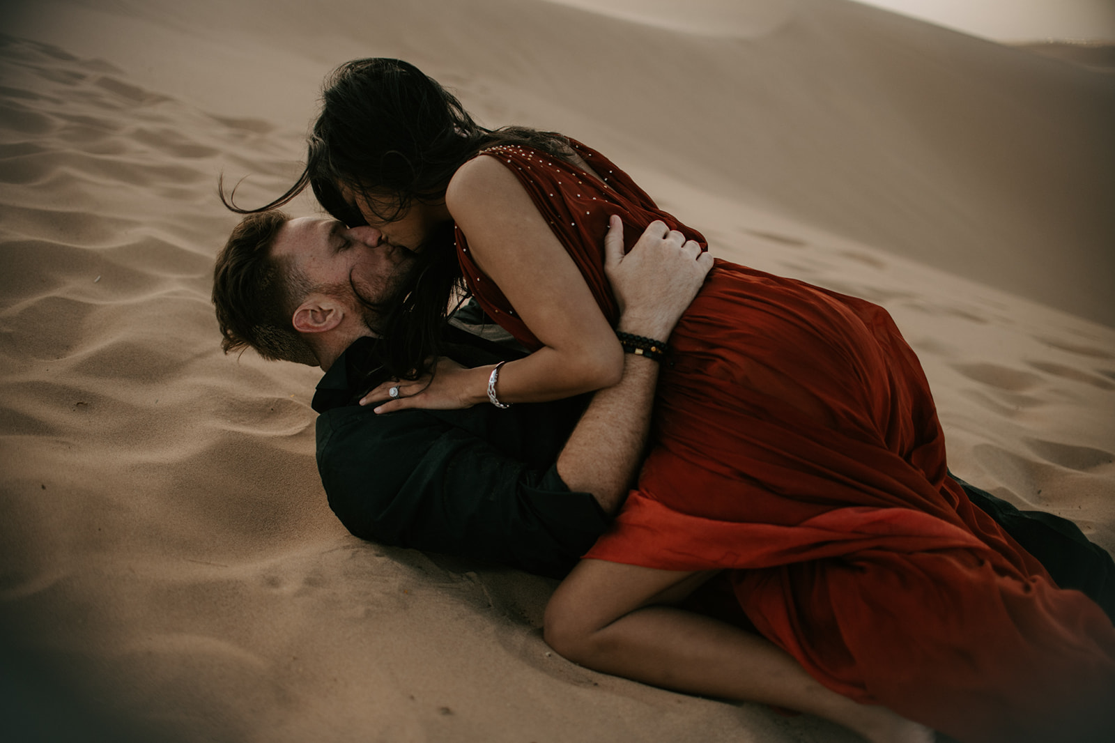 Couple laying together in the sand, sharing a kiss during a relaxed desert engagement moment

