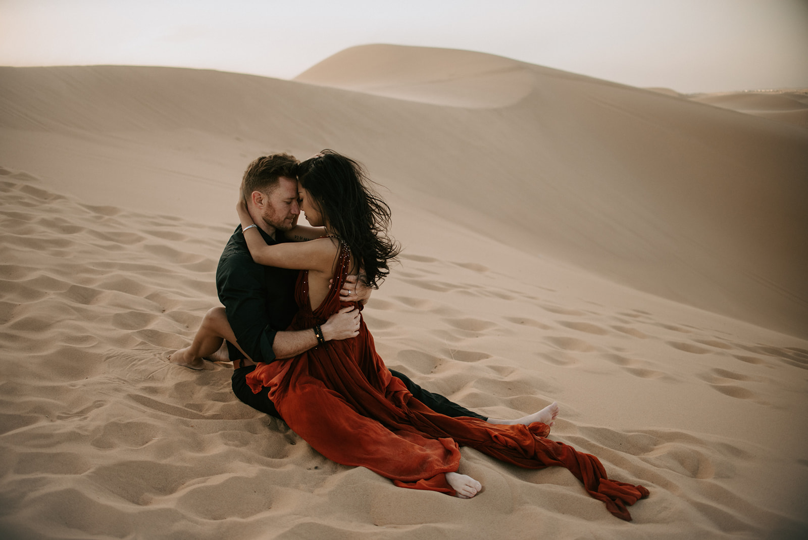 Couple sitting together in the sand, wrapped up in each other with flowing dress spread across the dunes
