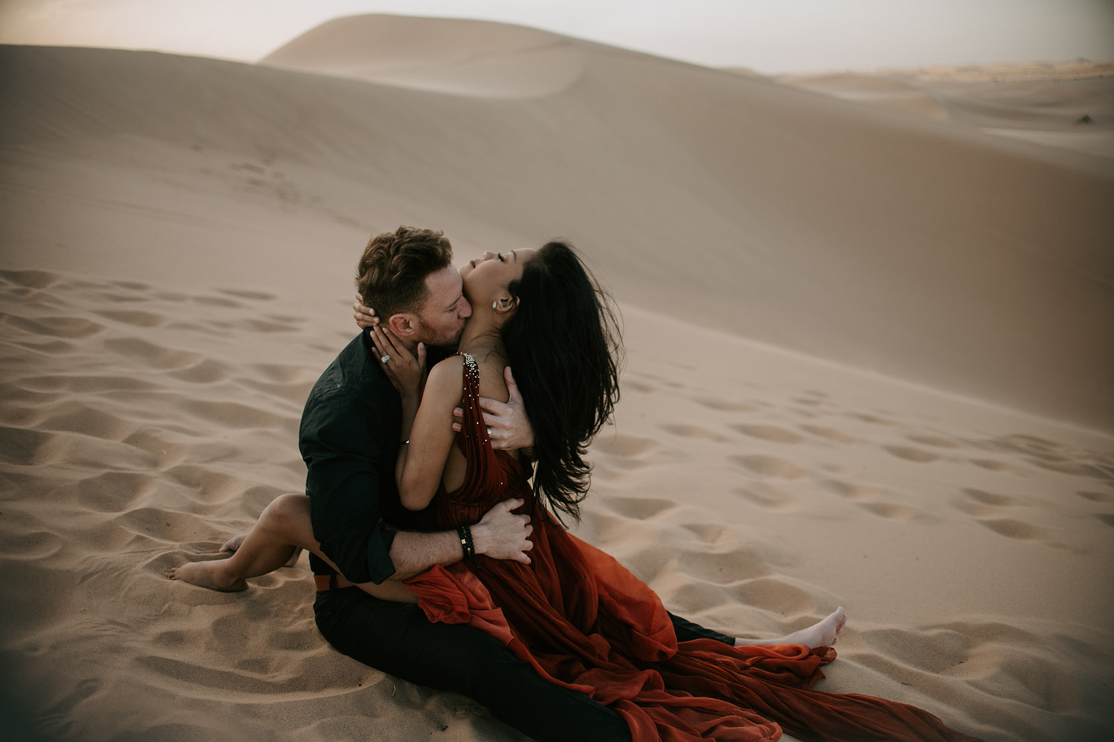 Couple sitting closely on the sand dunes, warm tones and flowing dress during a sand dunes photoshoot
