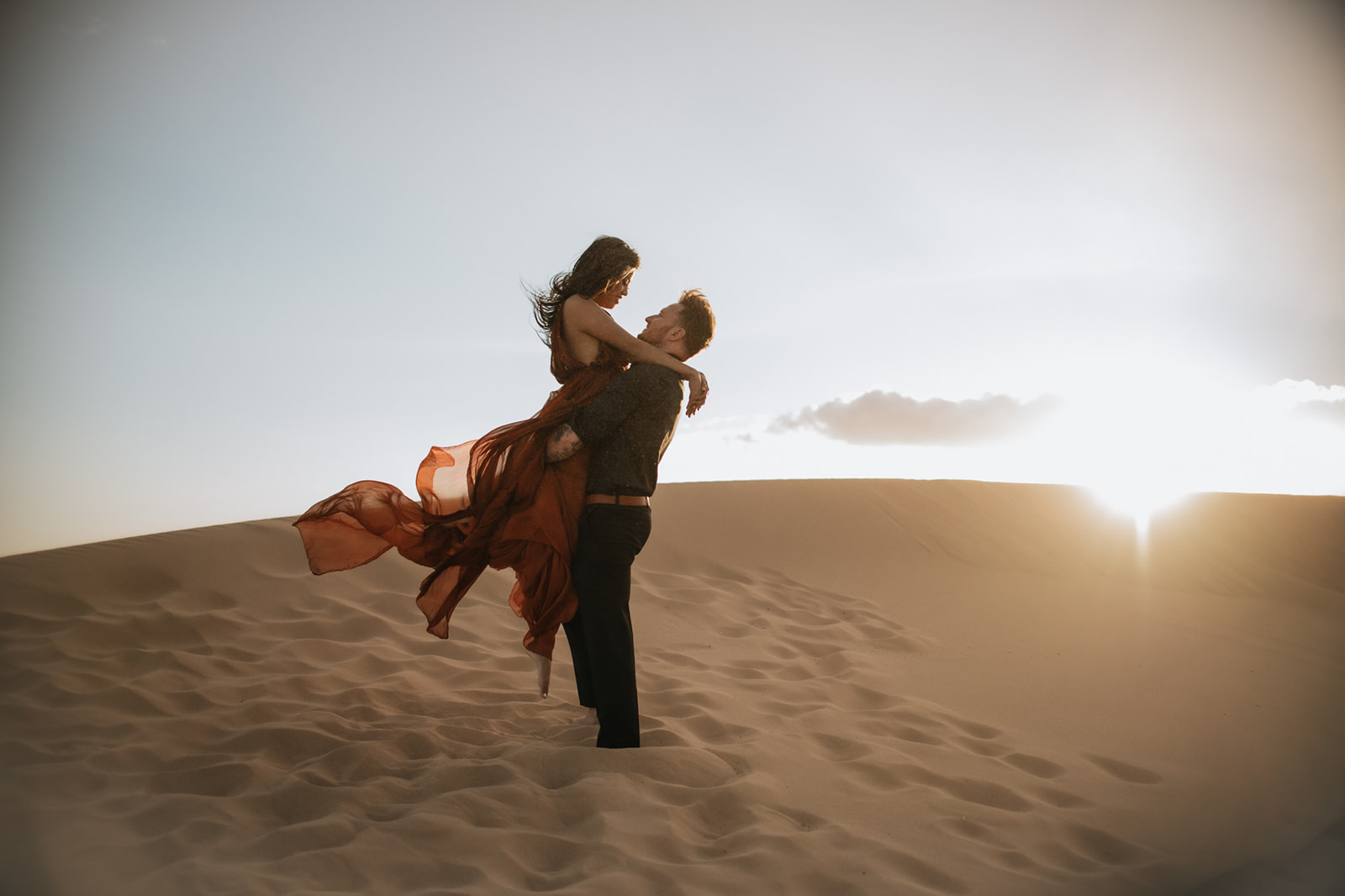 Couple lifting and holding each other on a dune at sunset, dress flowing in the wind during a sand dunes photoshoot
