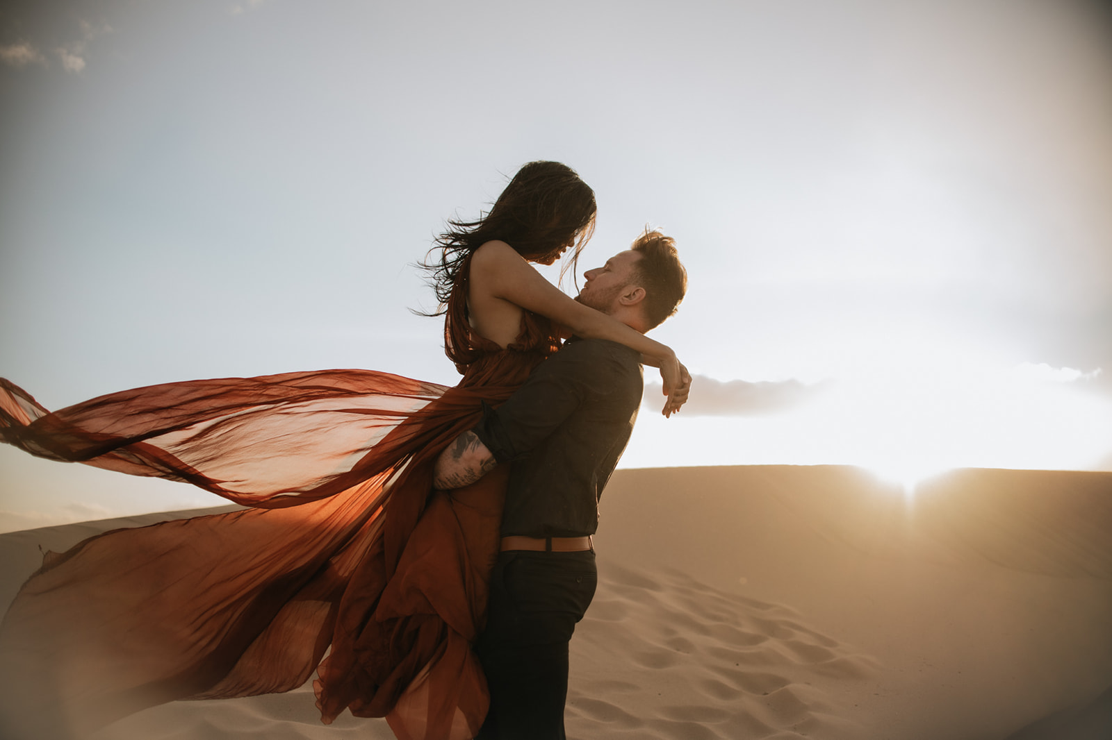 Couple embracing on a dune at sunset, wind catching the dress in a cinematic sand dunes photoshoot
