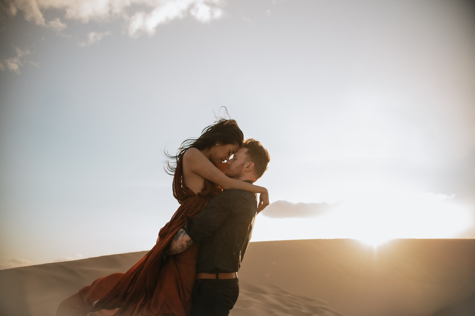 Couple embracing on a dune at sunset, warm light wrapping around them during a sand dunes photoshoot
