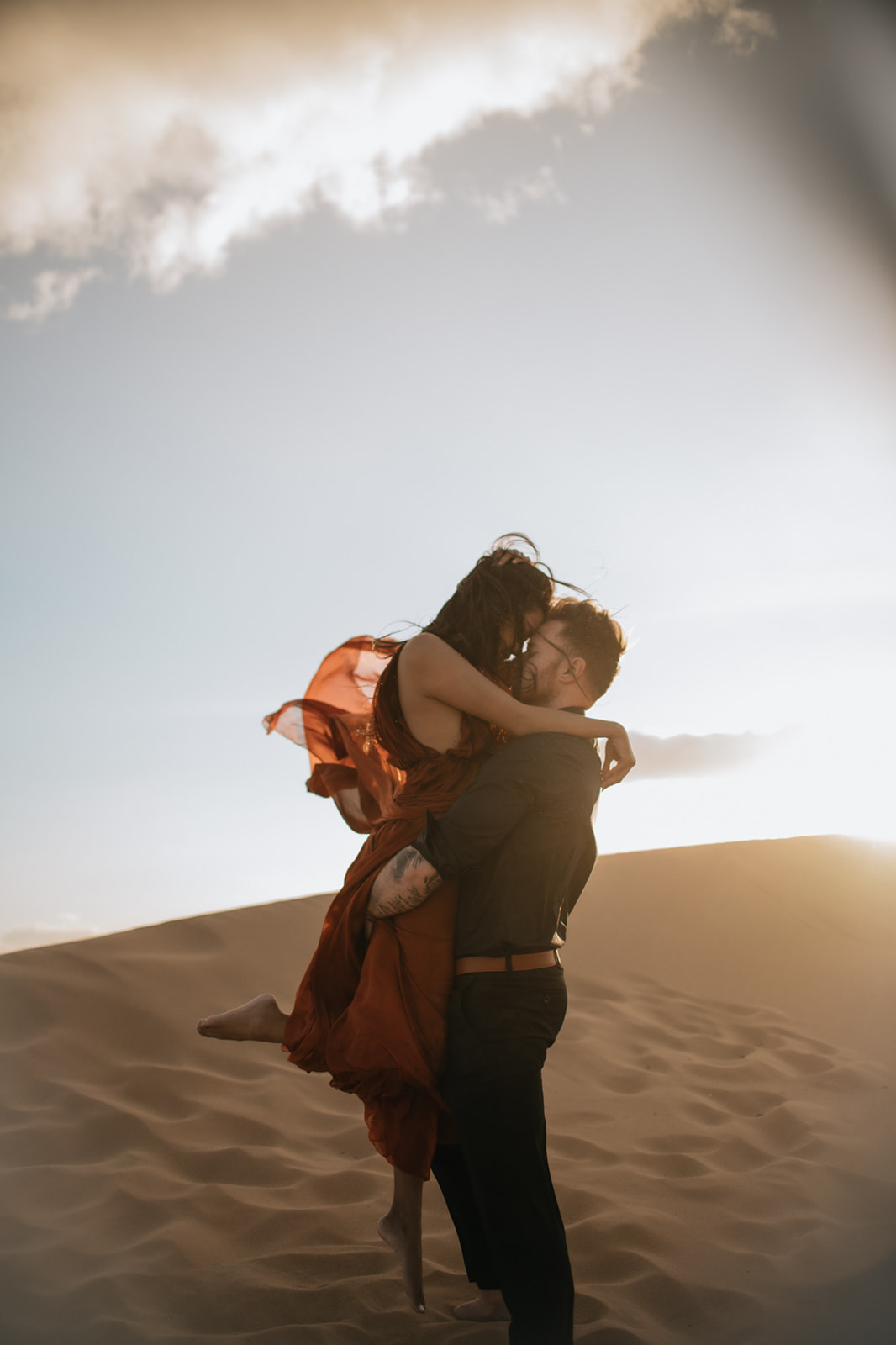 Couple embracing on a dune at sunset, backlit by warm light during a sand dunes photoshoot