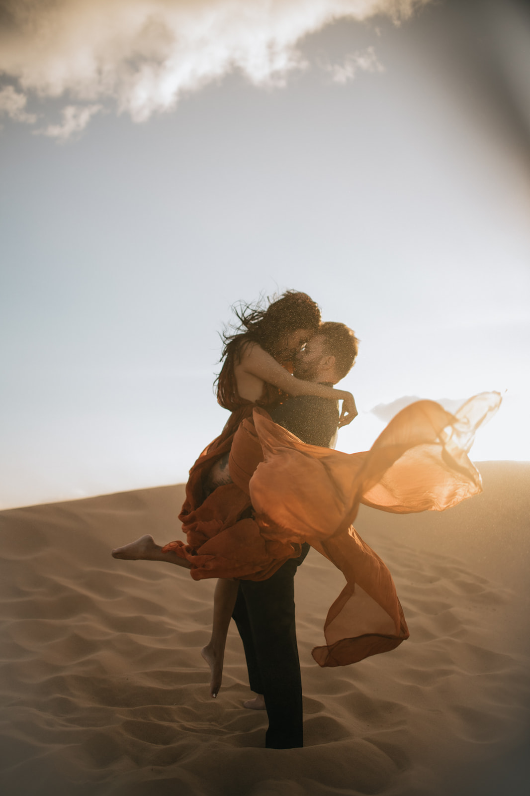 Couple embracing on a dune at golden hour, dress blowing dramatically in the wind during a sand dunes photoshoot
