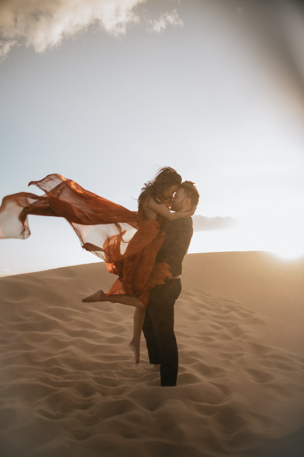 Couple lifting each other on a sunlit dune, red dress flowing dramatically in the wind during a sand dunes photoshoot

