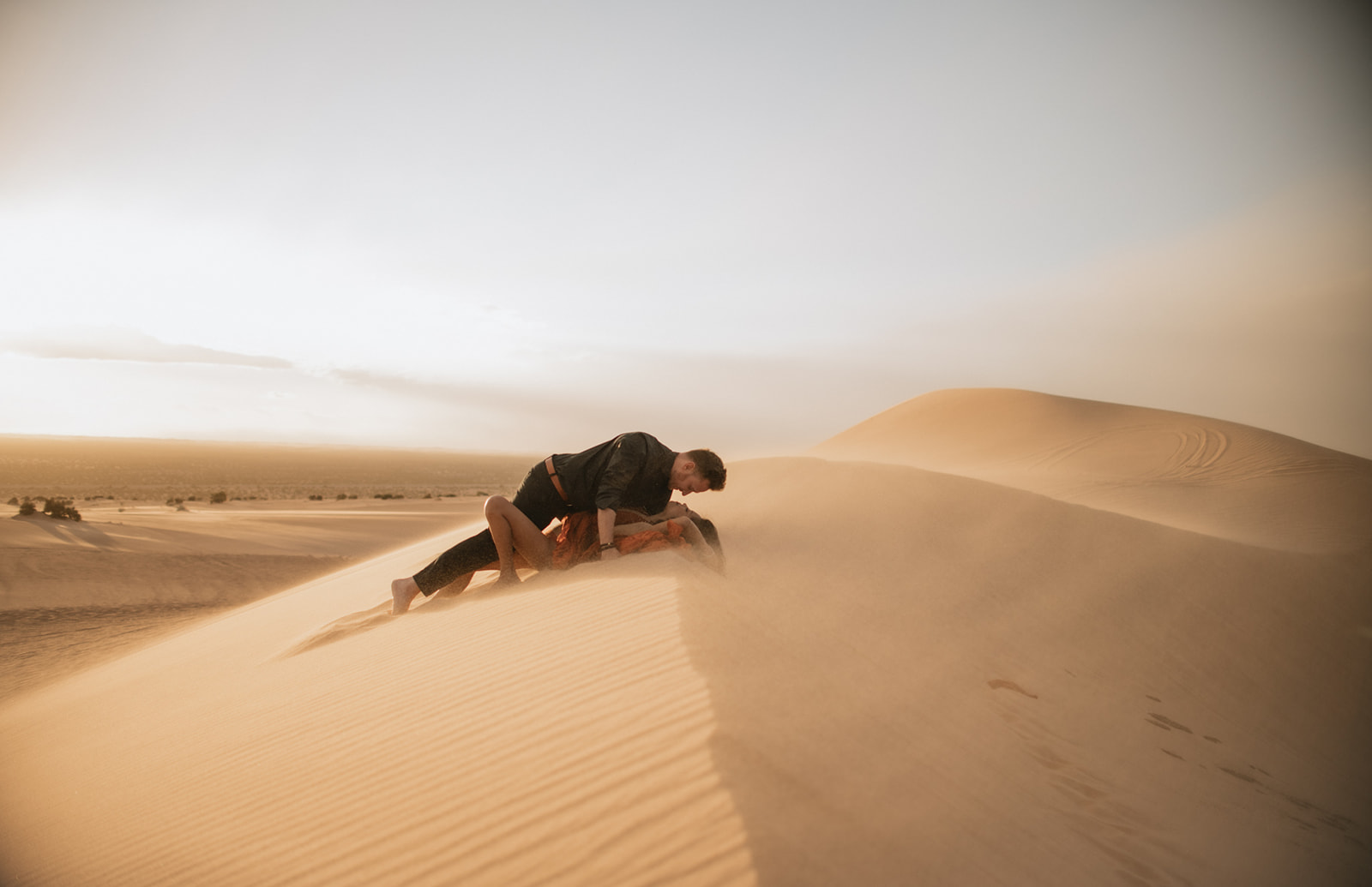 Wide shot of couple laying together on a dune, surrounded by soft golden sand during a sand dunes photoshoot
