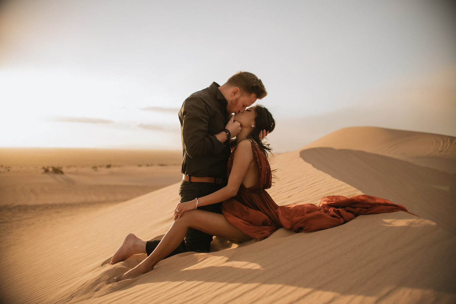 Couple sitting together on a dune at golden hour, sharing a kiss with soft desert light
