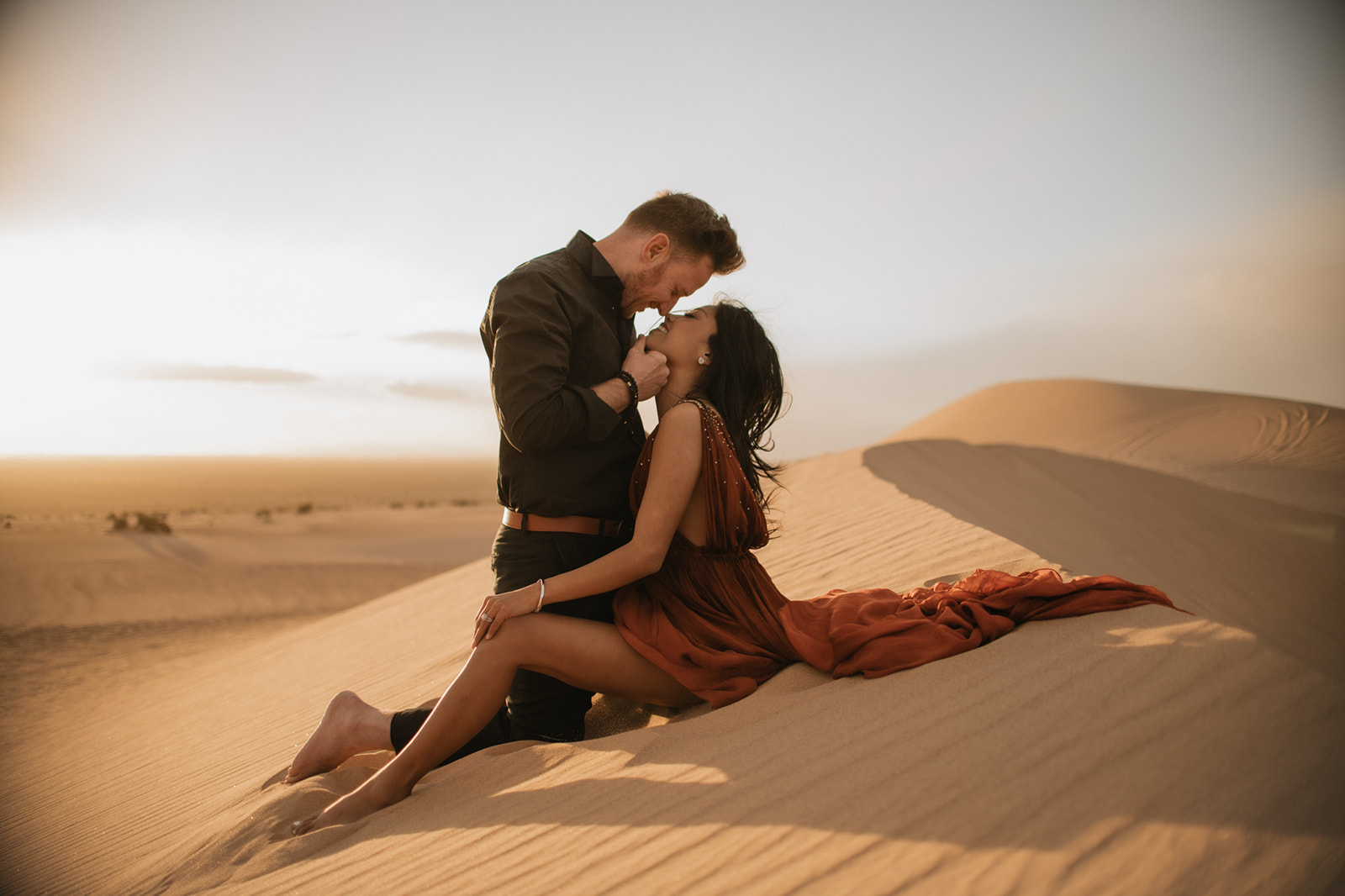 Couple leaning into each other on a sunlit dune, soft light and movement in her dress during a sand dunes photoshoot
