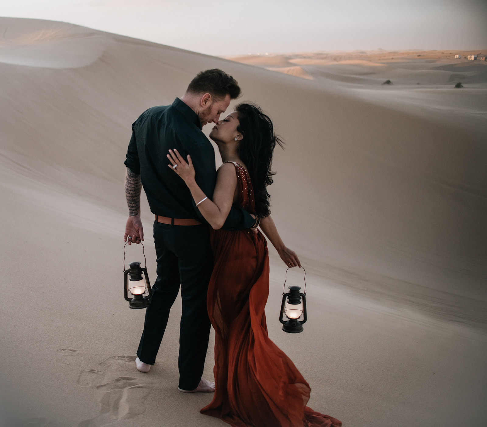 Couple walking along desert dunes holding lanterns, red dress trailing behind during a sand dunes photoshoot
