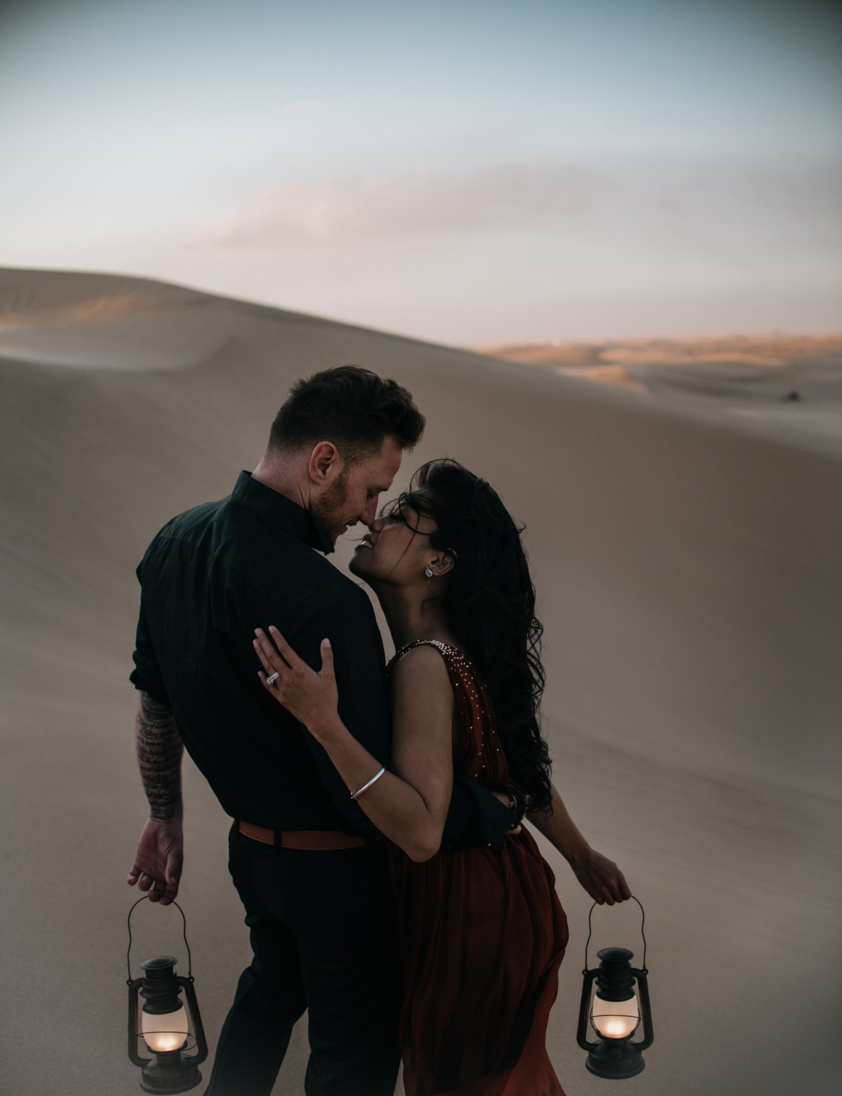 Couple walking through desert dunes holding lanterns, soft evening light and wind creating an intimate sand dunes photoshoot moment
