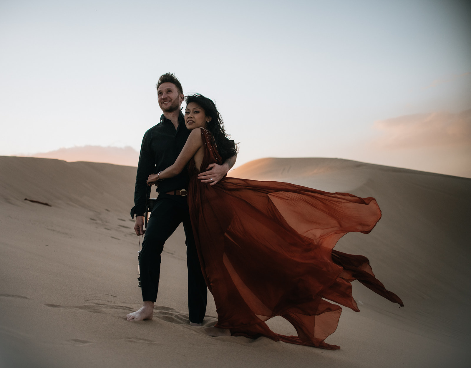 Couple walking away together across soft dunes at sunset, footprints trailing behind during a sand dunes photoshoot
