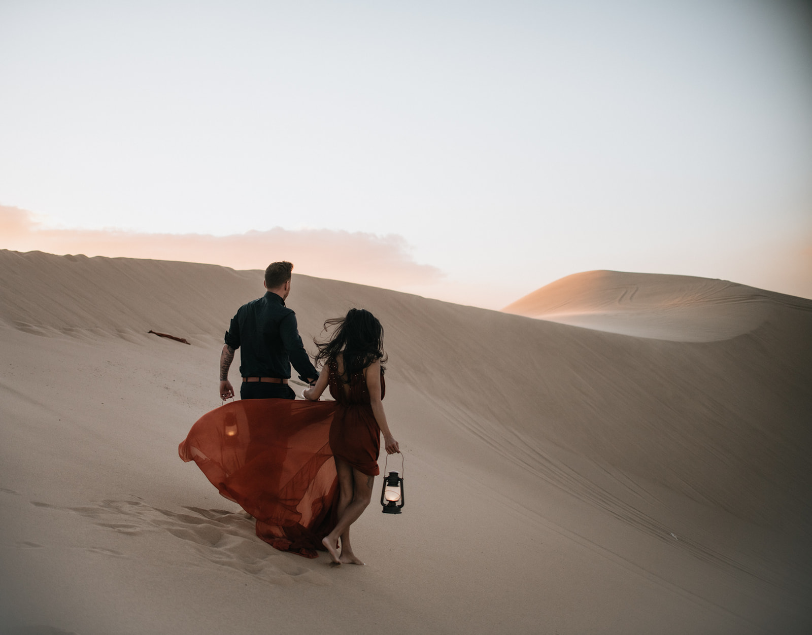 Couple walking hand in hand across desert dunes carrying lanterns, cinematic and windswept sand dunes photoshoot