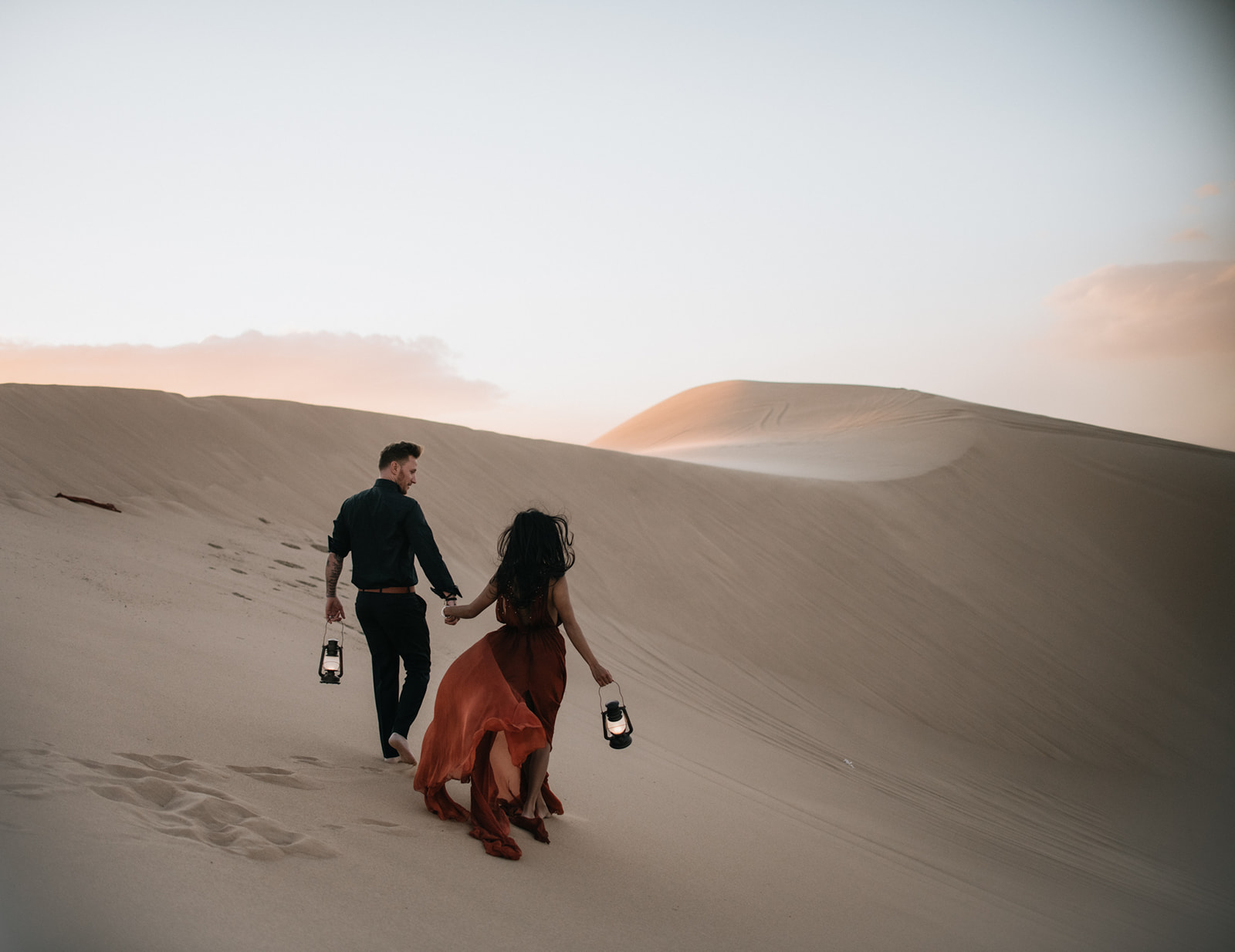 Wide shot of couple standing on a dune at golden hour, surrounded by soft light and rolling sand dunes
