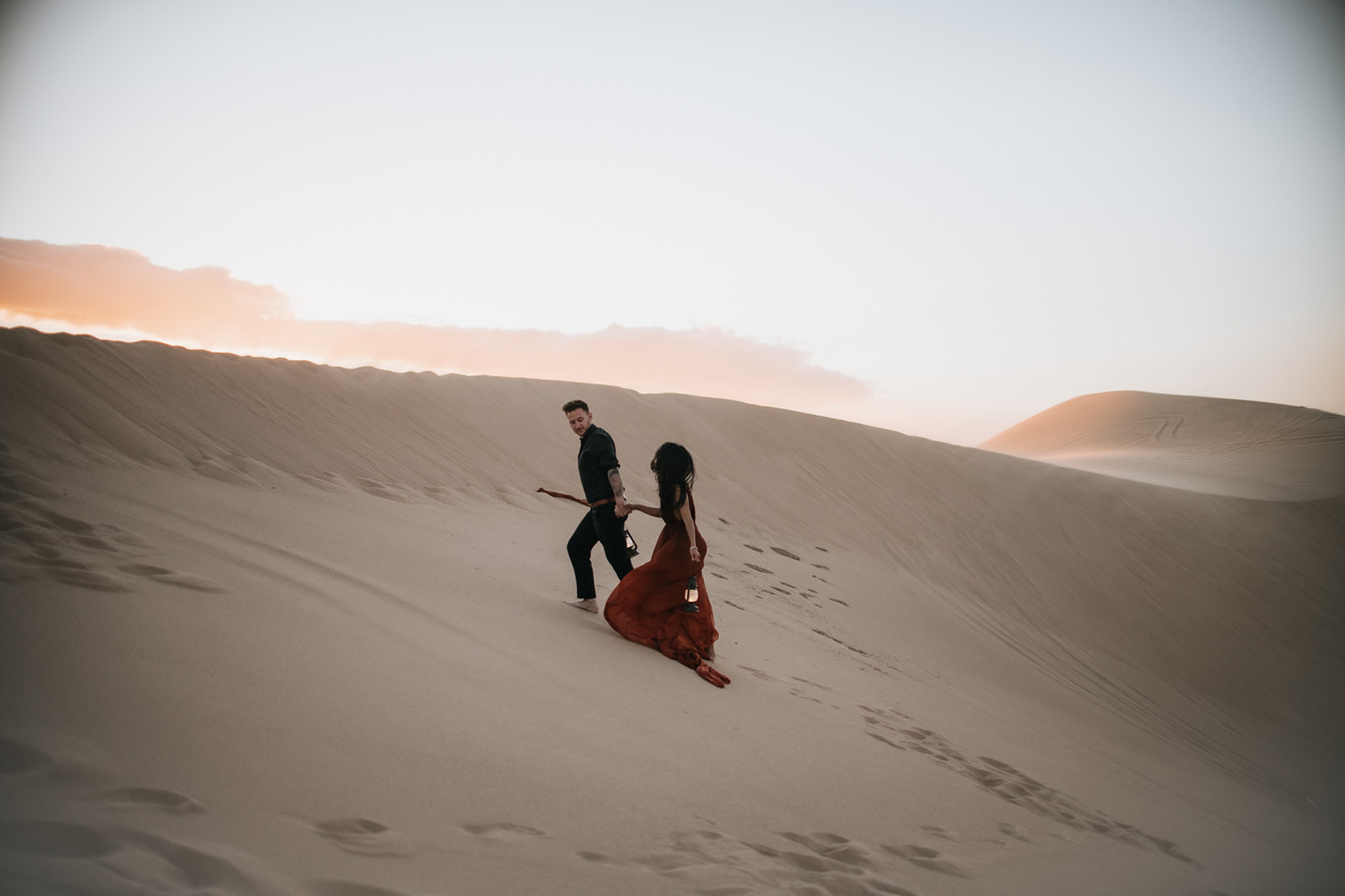 Couple walking hand in hand up a dune holding lanterns, footprints trailing behind in the sand dunes photoshoot
