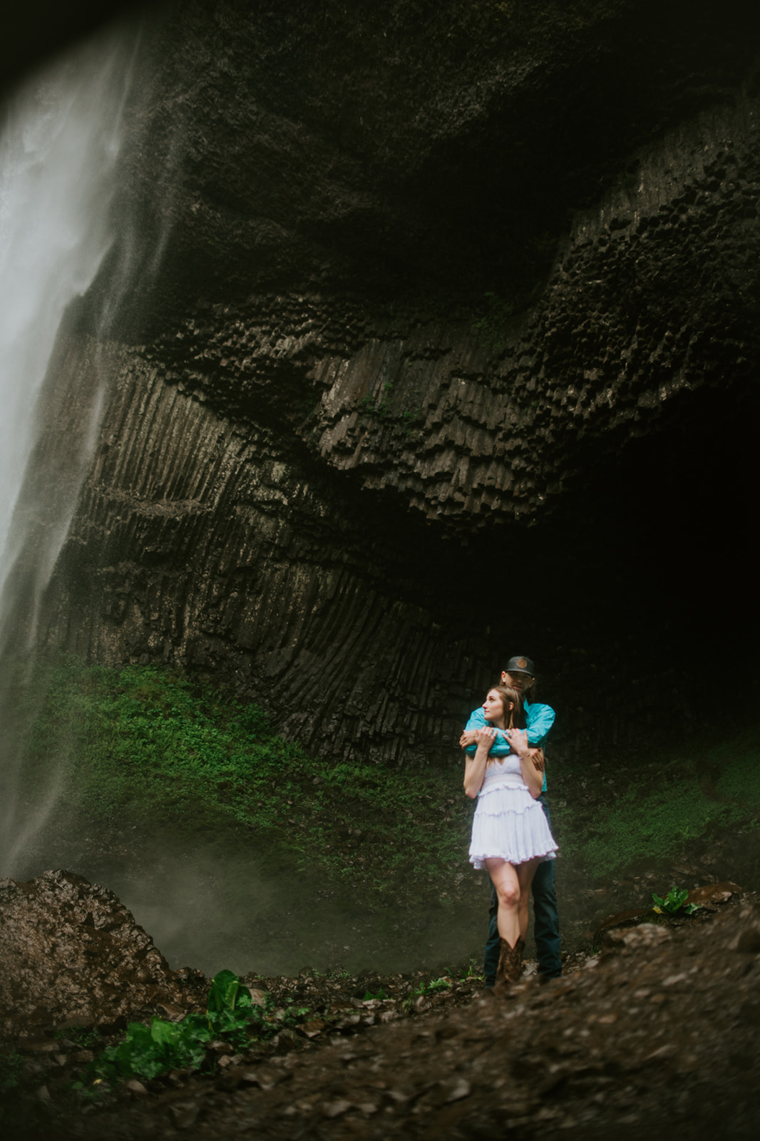 Couple hugging on wooden bridge with waterfall behind them, intimate outdoor engagement photos