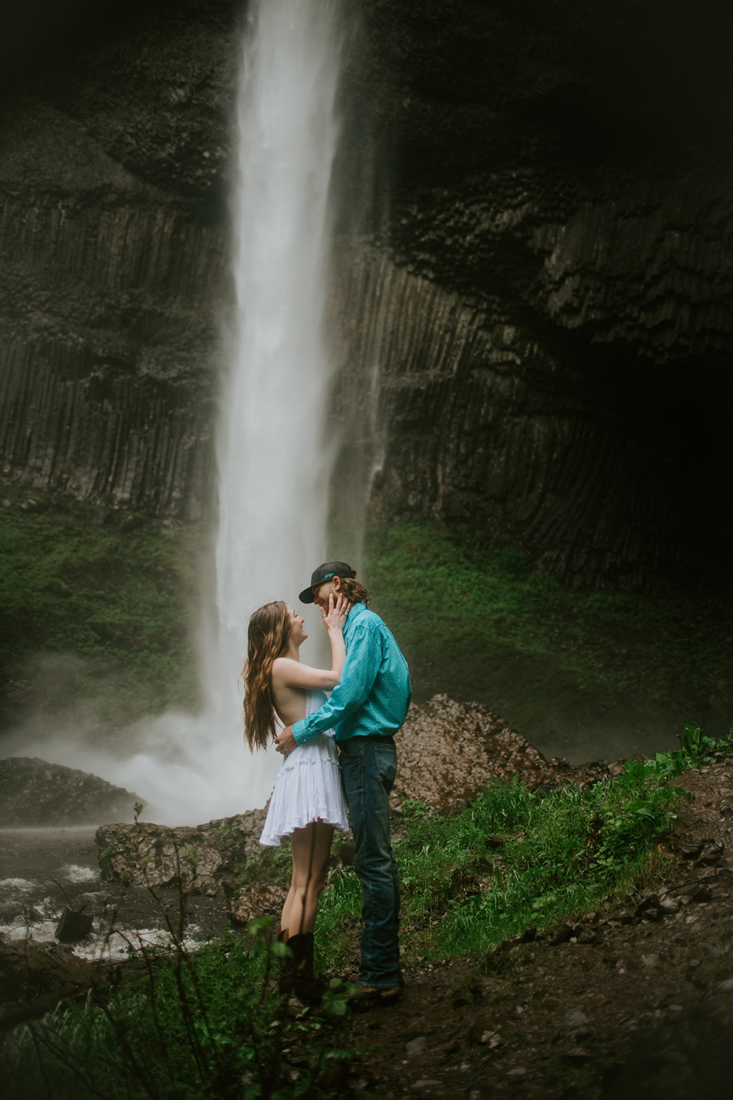Couple standing close together in front of a tall waterfall, intimate and moody outdoor engagement photos