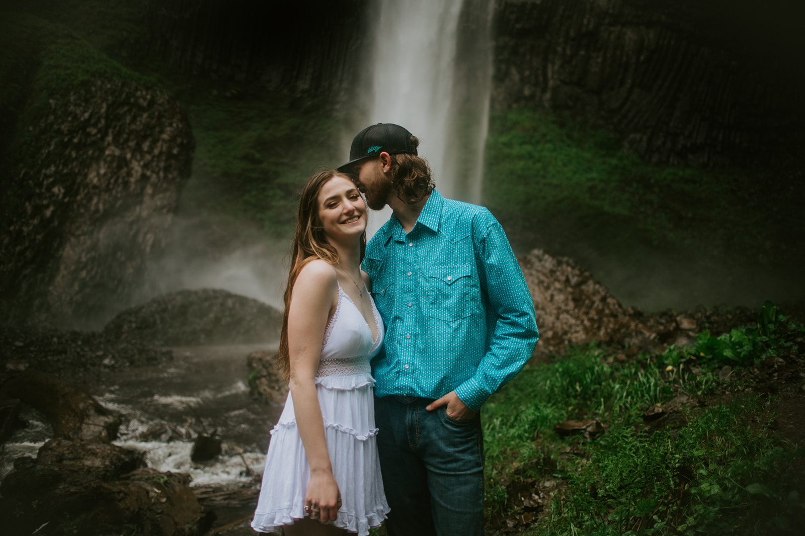 Couple smiling together in front of waterfall with lush greenery during outdoor engagement photos