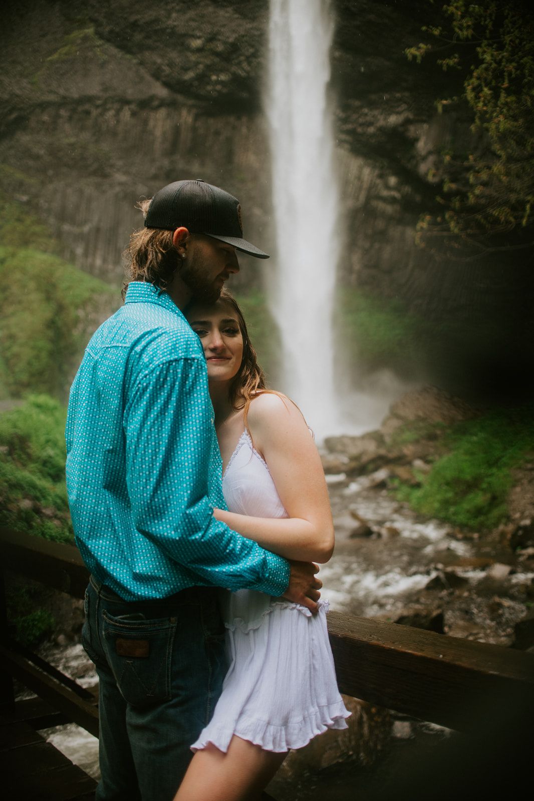 Couple hugging along a wooden bridge with waterfall behind them during outdoor engagement photos