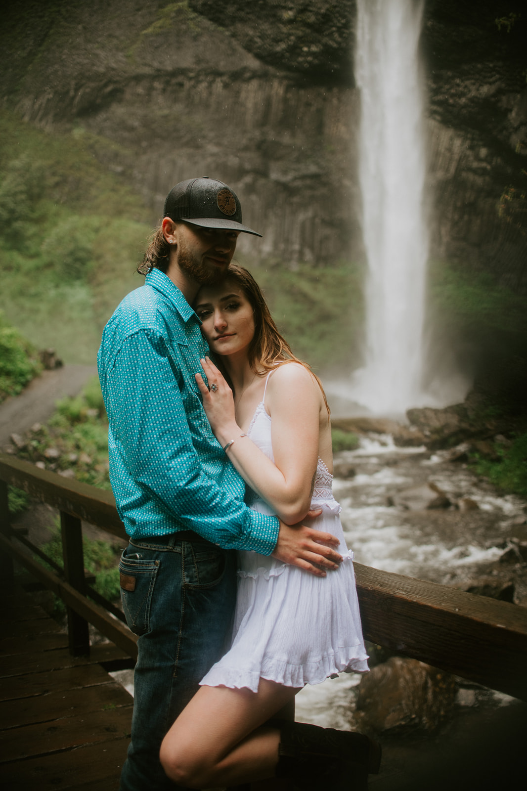 Couple embracing on a wooden railing with waterfall behind them during outdoor engagement photos in Oregon