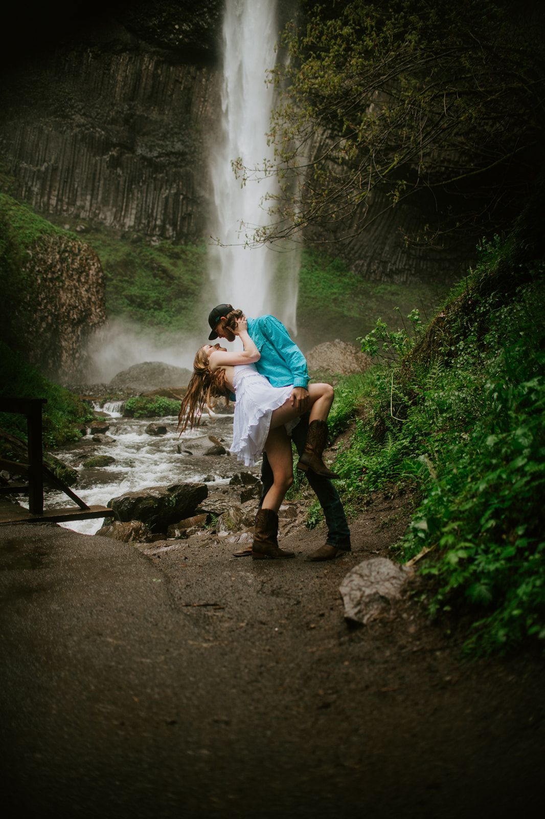 Couple dipping for a kiss on a forest path with waterfall in the background during outdoor engagement photos