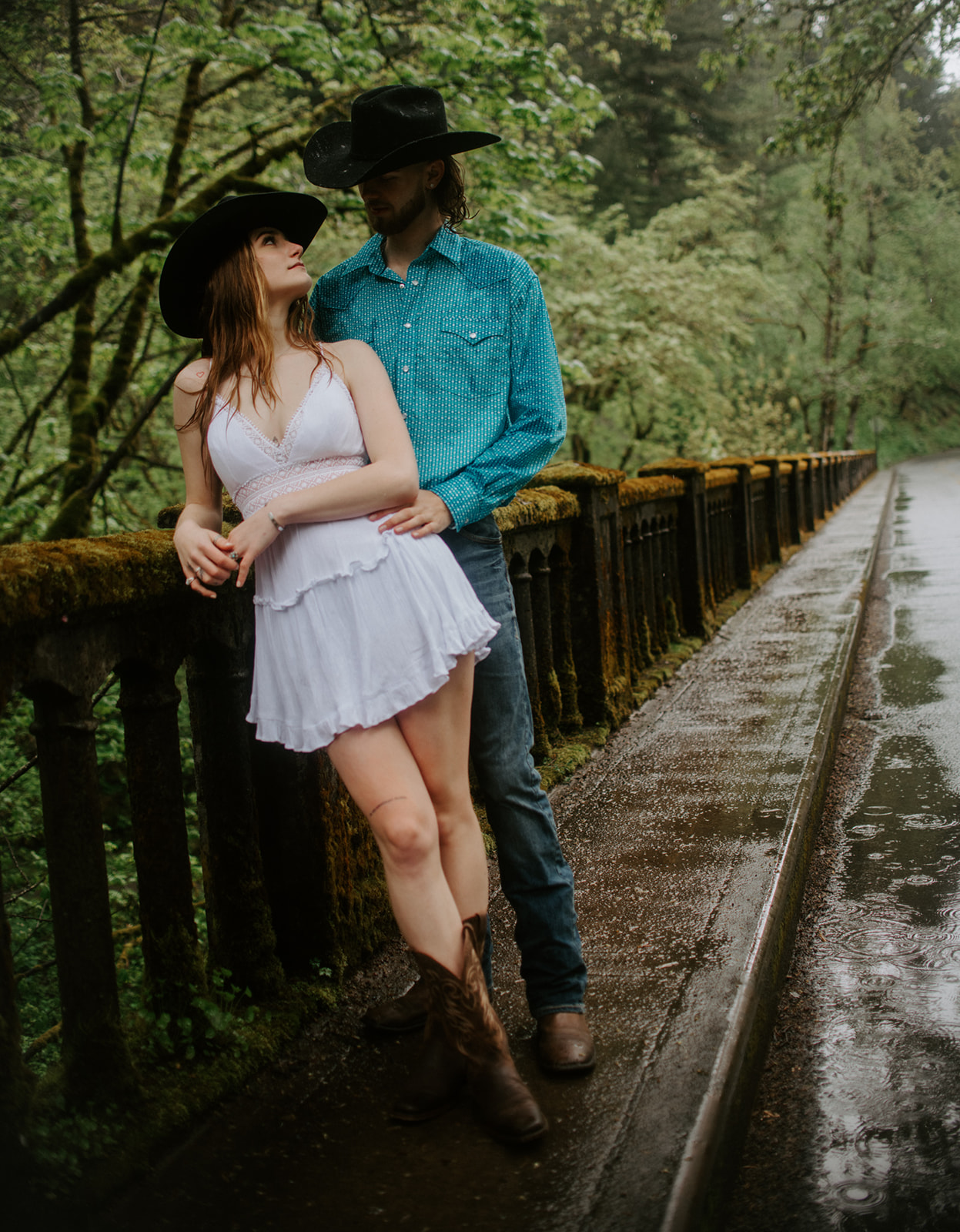 Couple leaning against mossy railing on a forest bridge, relaxed and candid outdoor engagement photos
