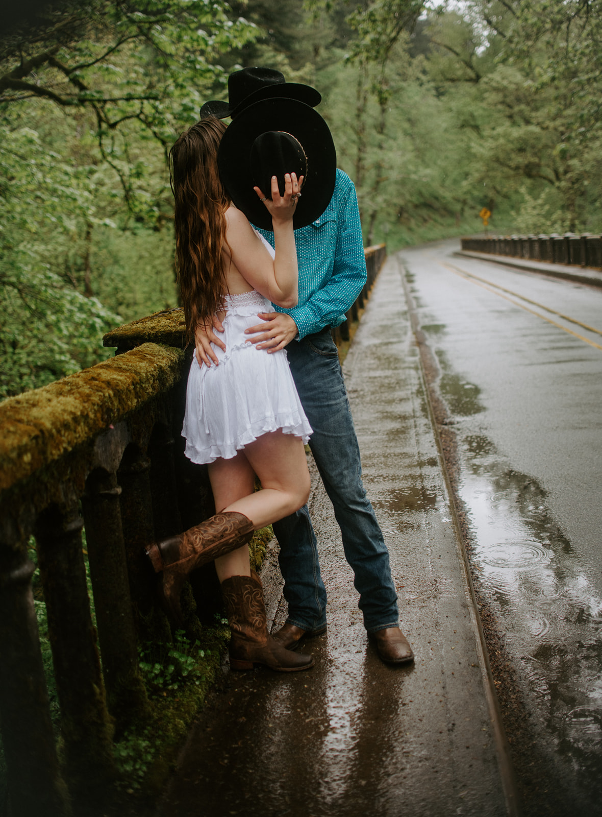Couple sharing a kiss on a rainy road with cowboy boots and hats, playful and candid engagement moment