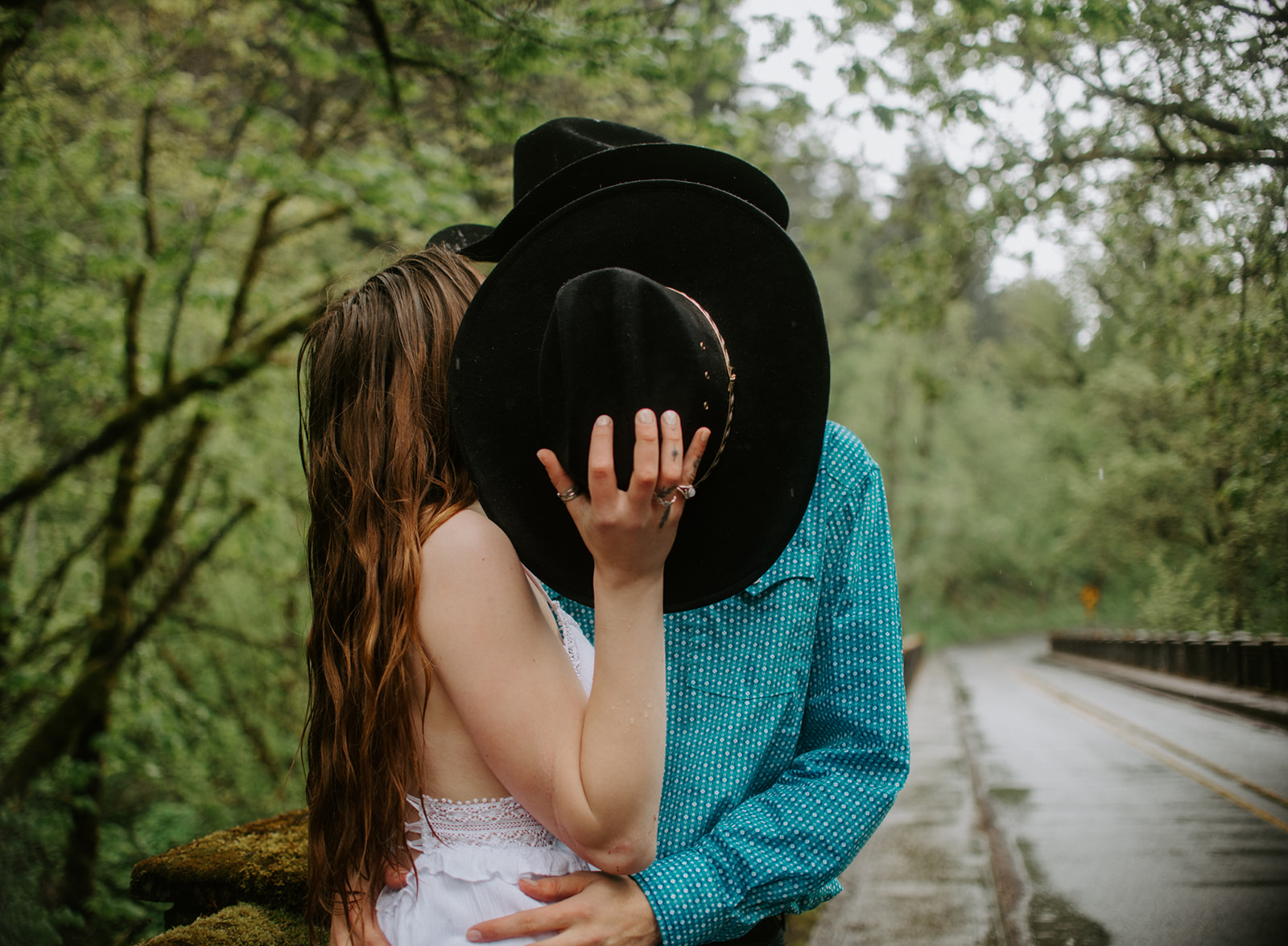 Couple sharing a kiss under cowboy hats on a rainy forest road, playful engagement photo
