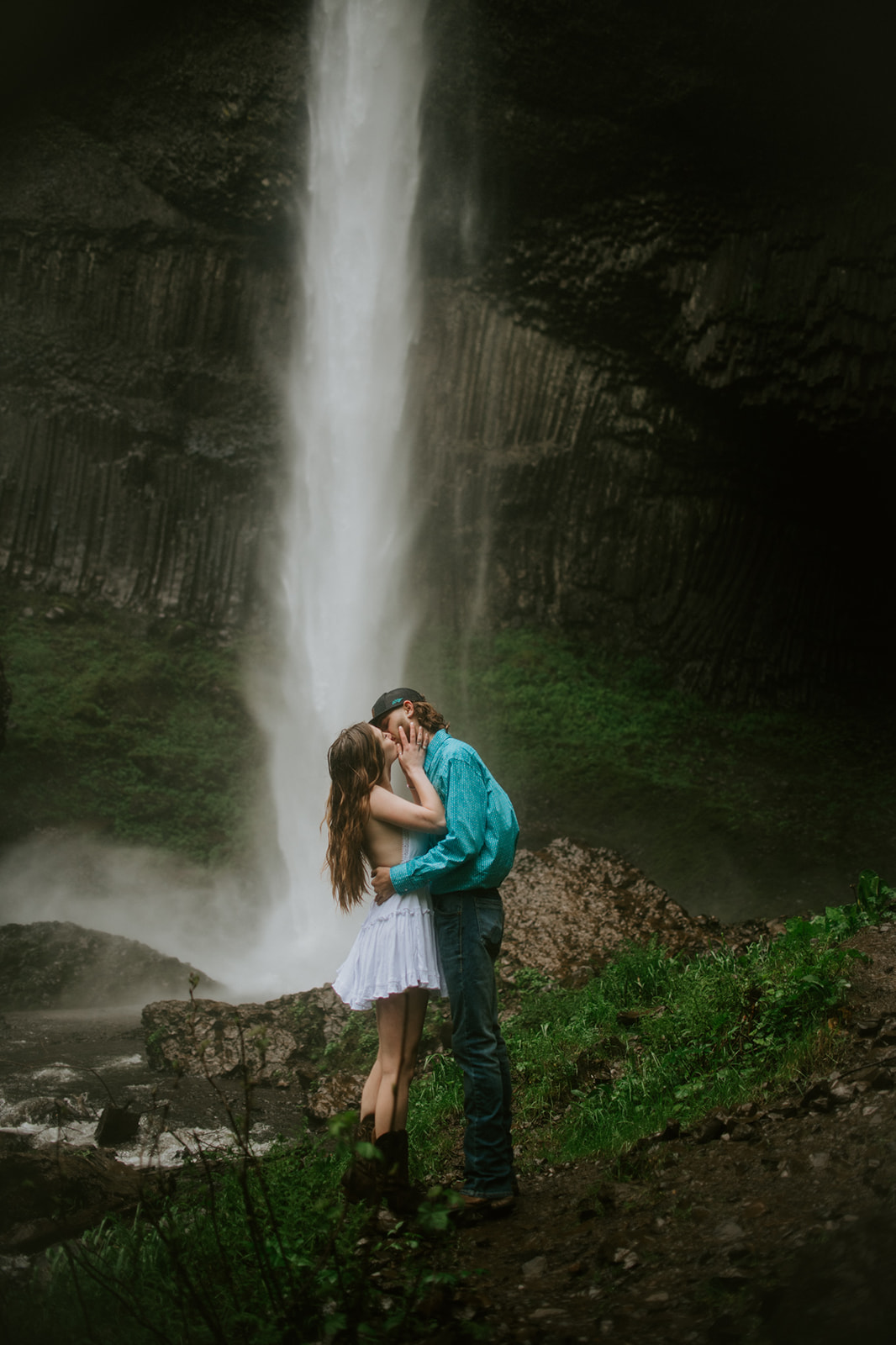 Couple kissing in front of a dramatic waterfall during a misty, adventurous engagement photoshoot