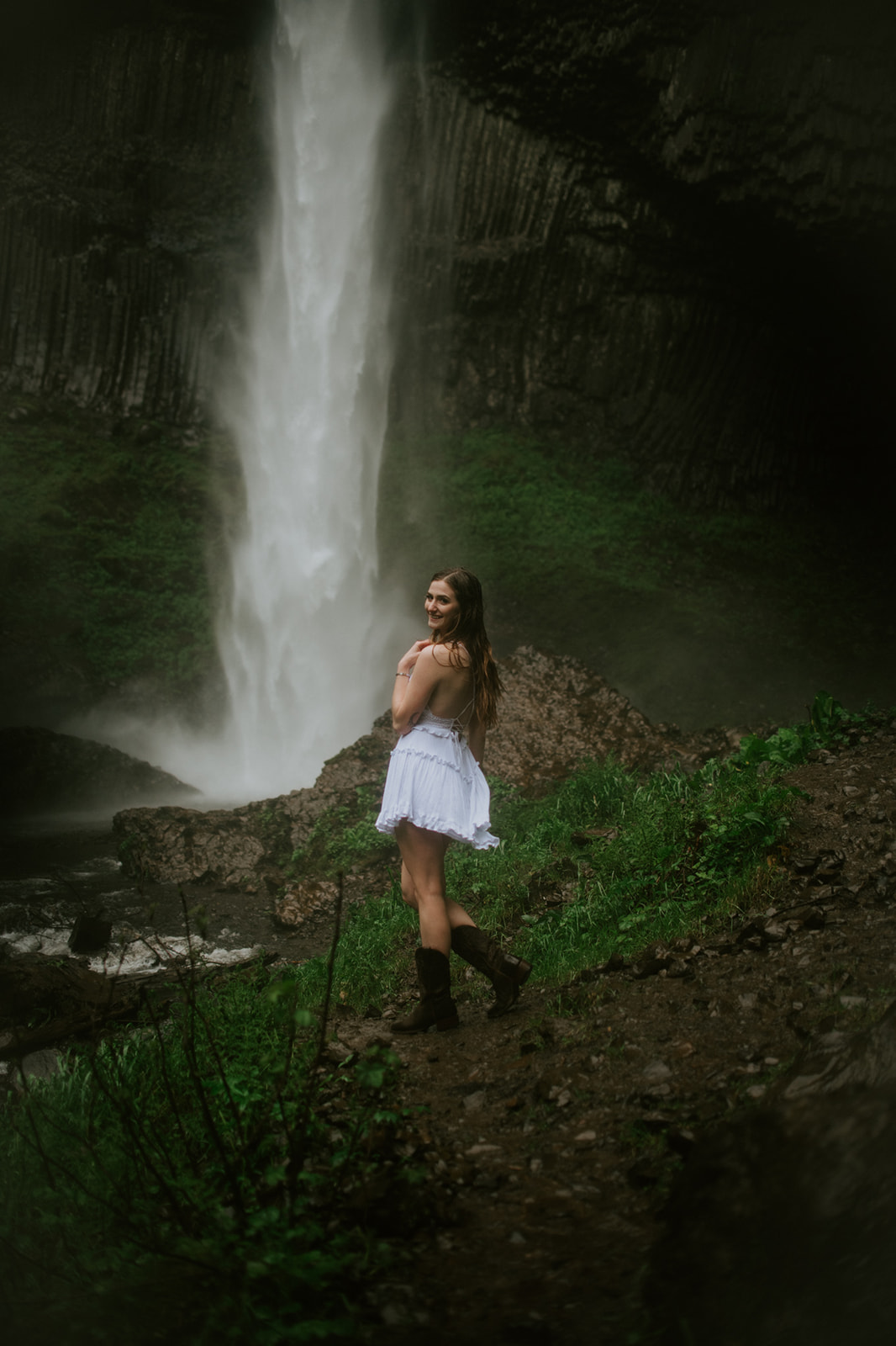 Bride-to-be looking back and smiling while standing near a misty waterfall on a lush green trail