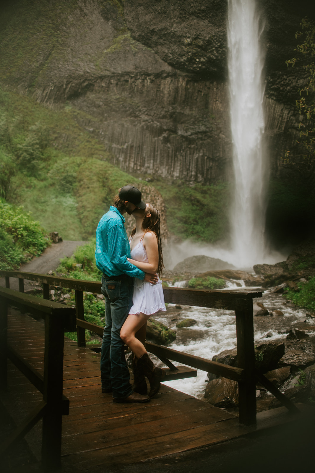 Couple kissing on a wooden bridge in front of a powerful waterfall surrounded by lush greenery during an engagement photoshoot