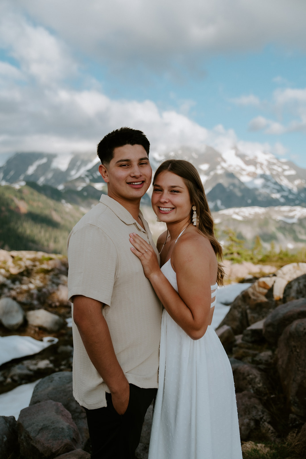 Smiling couple posing together in the mountains with rocky terrain and snow-capped peaks behind them