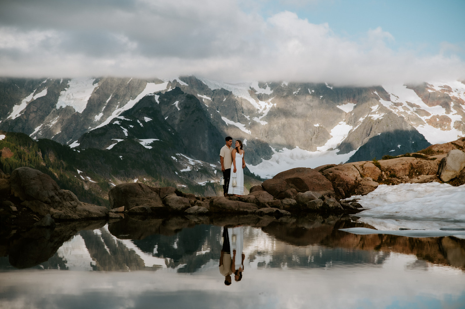 Scenic engagement photoshoot with couple standing on rocky ledge reflected in an alpine lake with snowy mountains behind them